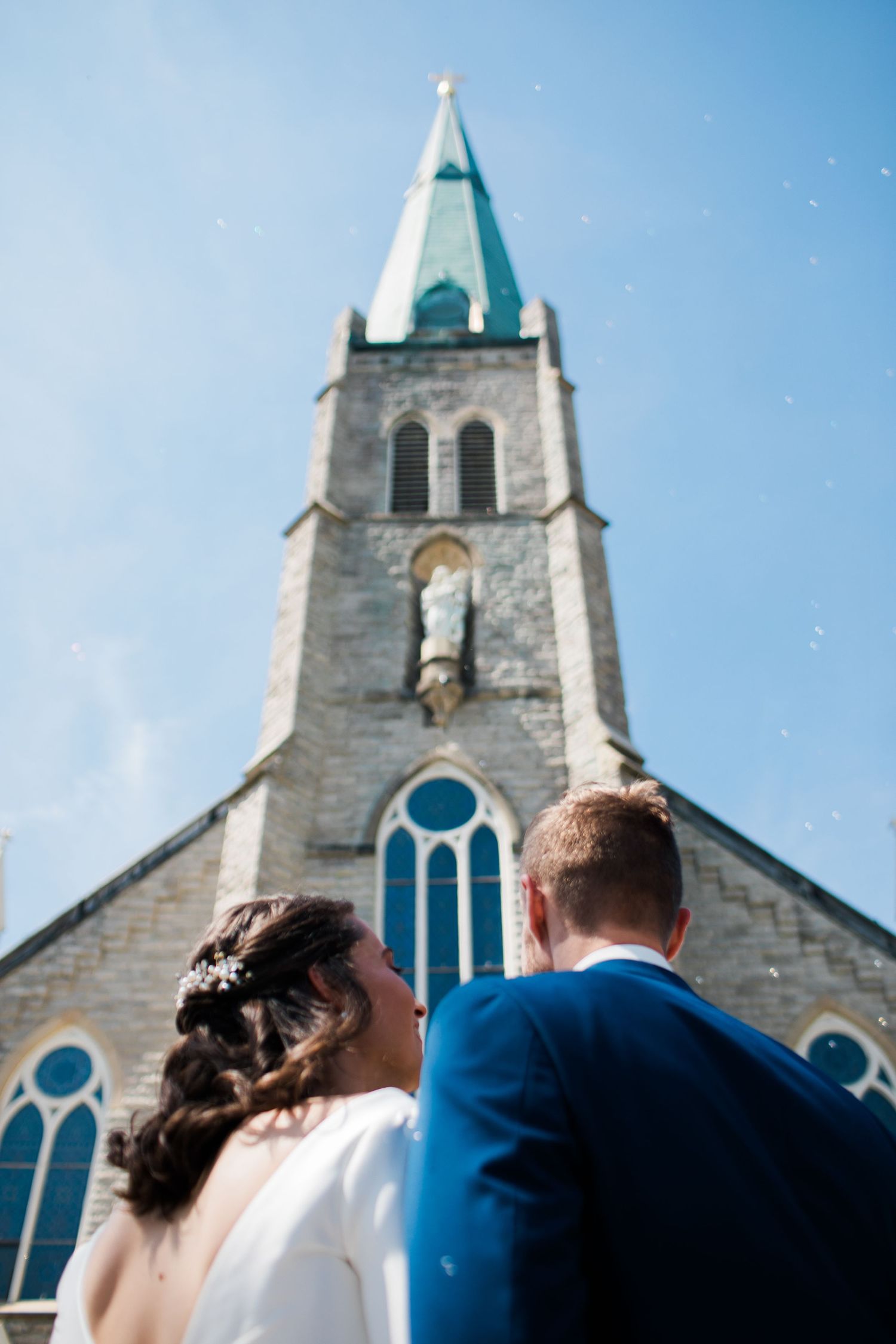 Bride and groom stand before a stone church with a tall steeple on their wedding day.