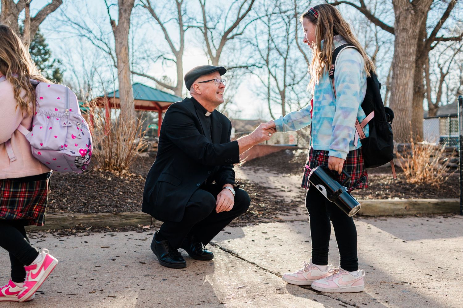 A priest kneels to greet a young girl with a handshake outdoors near a school on a winter day.