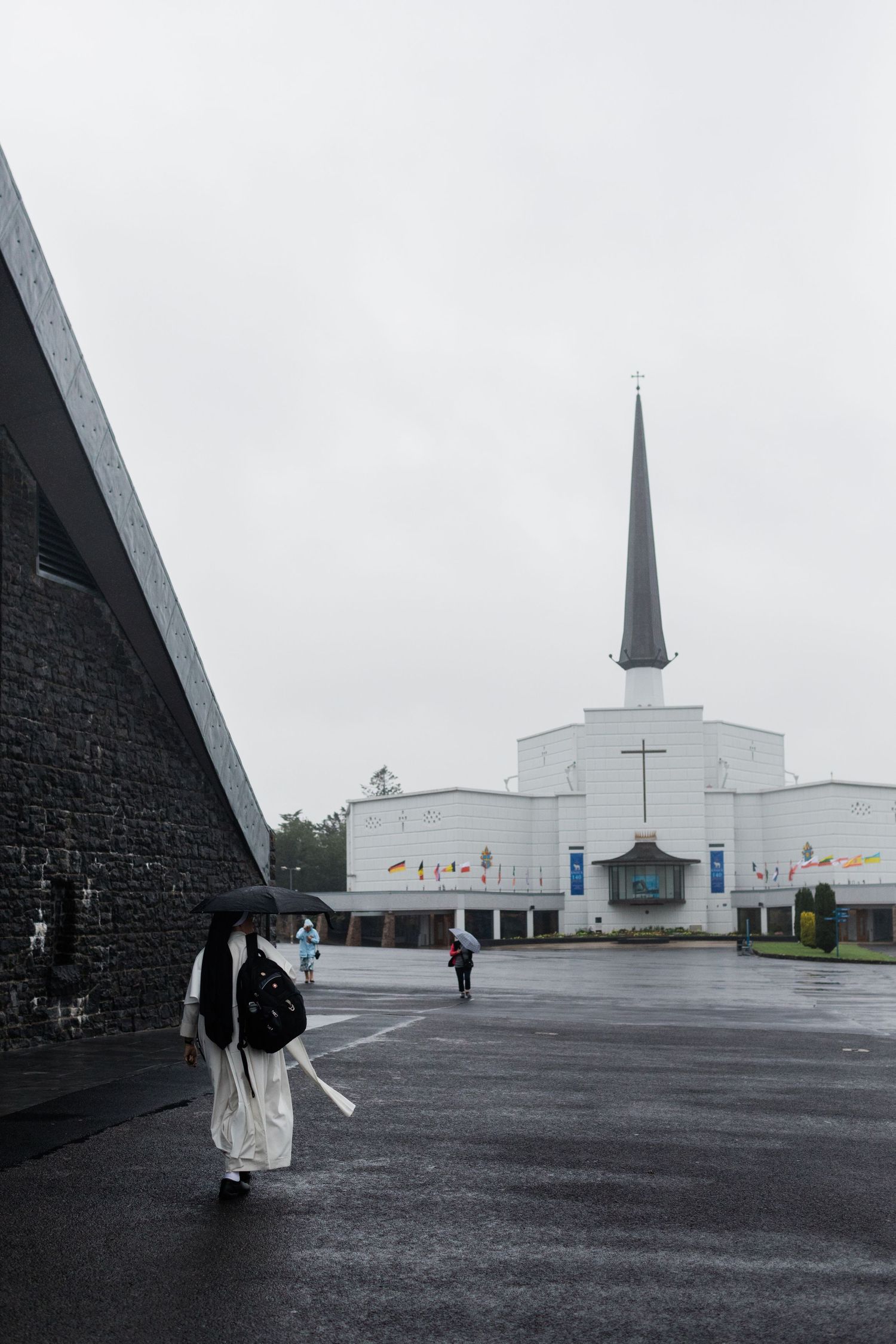 Catholic nun with umbrella walks toward a white modern church with a tall steeple on a rainy day.