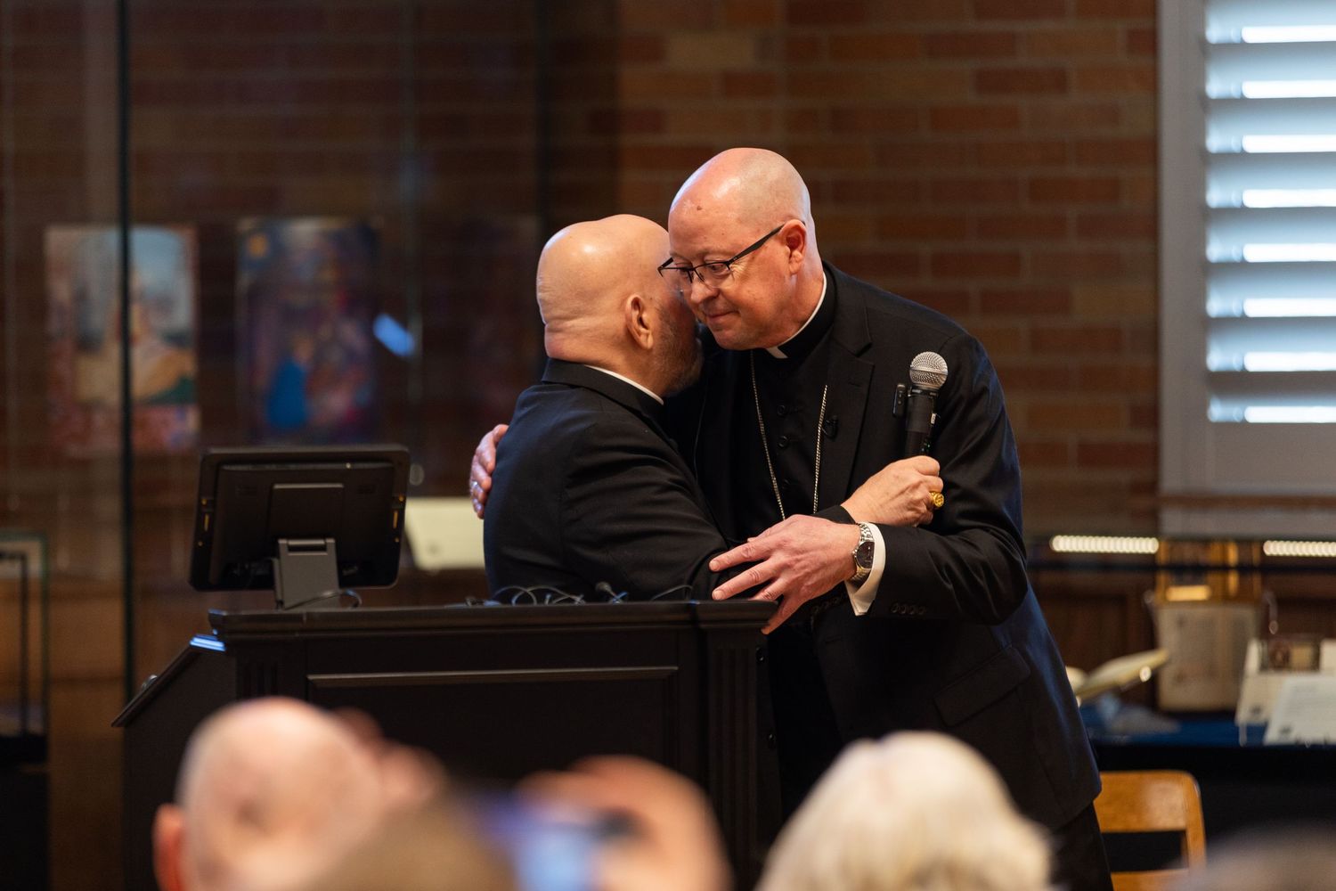 Two Catholic clergy members share an embrace during a formal church gathering, one holding a microphone.