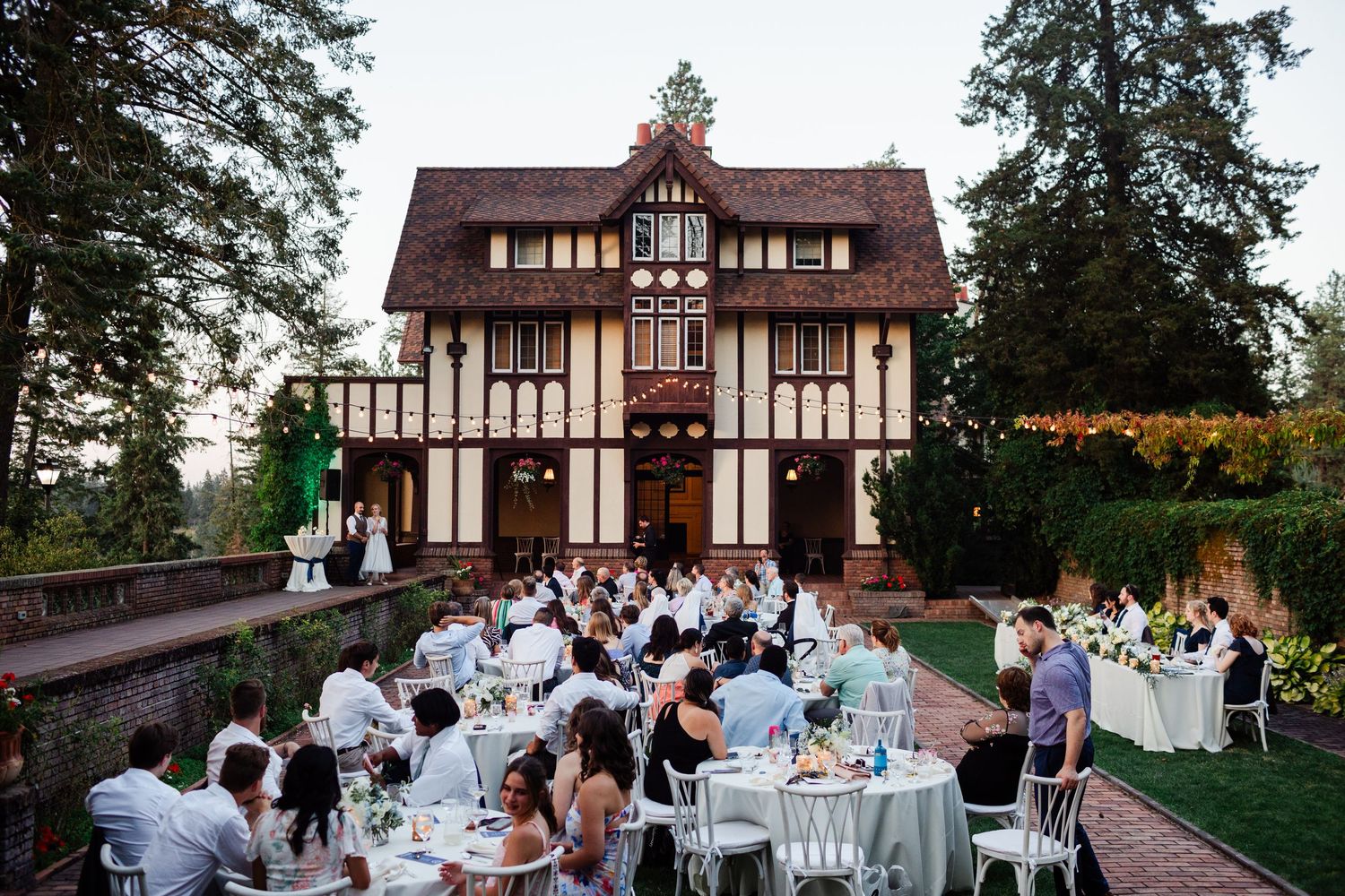 Evening outdoor wedding reception with guests seated at white tables in front of a Tudor mansion.