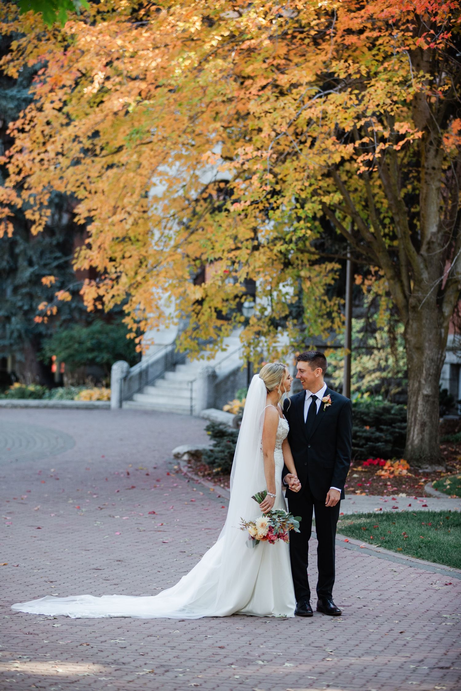 Bride and groom holding hands on brick path surrounded by vibrant autumn foliage during fall wedding portrait.
