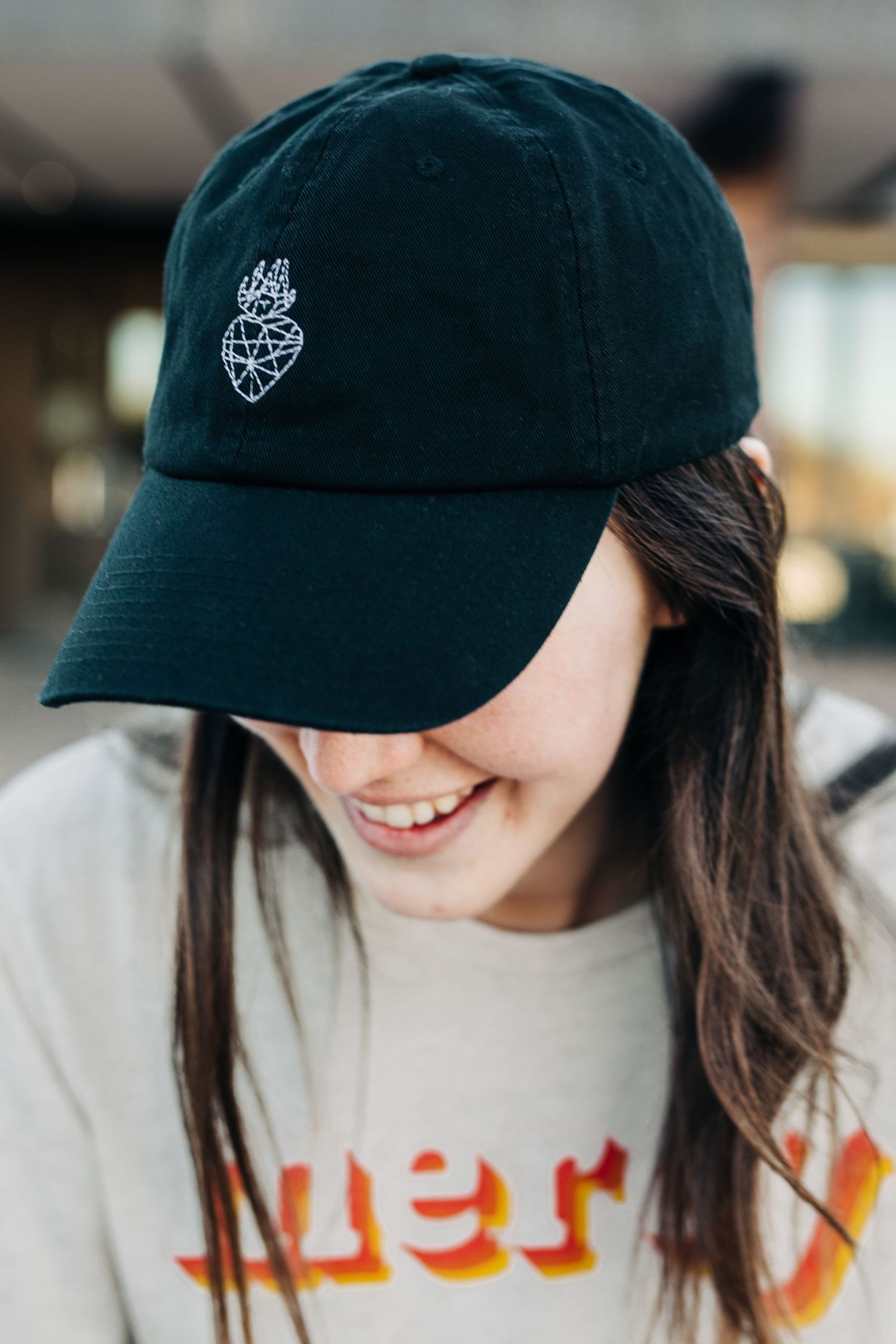 A black baseball cap with embroidered logo worn by someone in a beige sweatshirt with orange lettering.