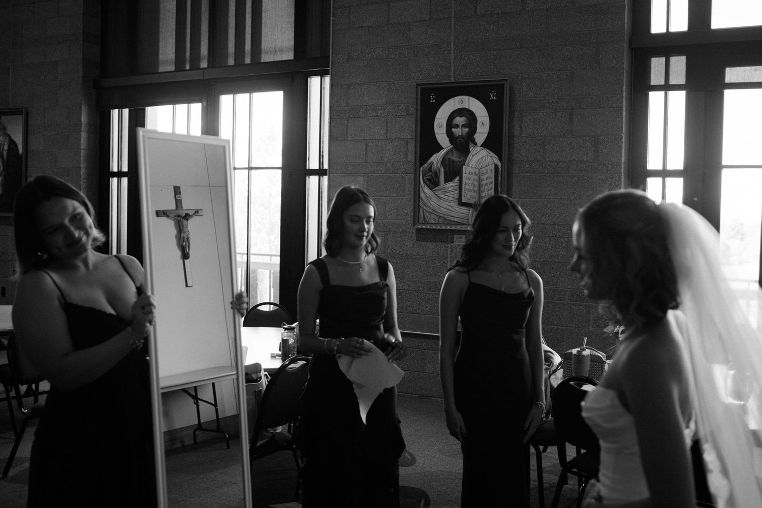 Black and white photo of bridesmaids and bride gathering near a crucifix painting in a church before a wedding ceremony.