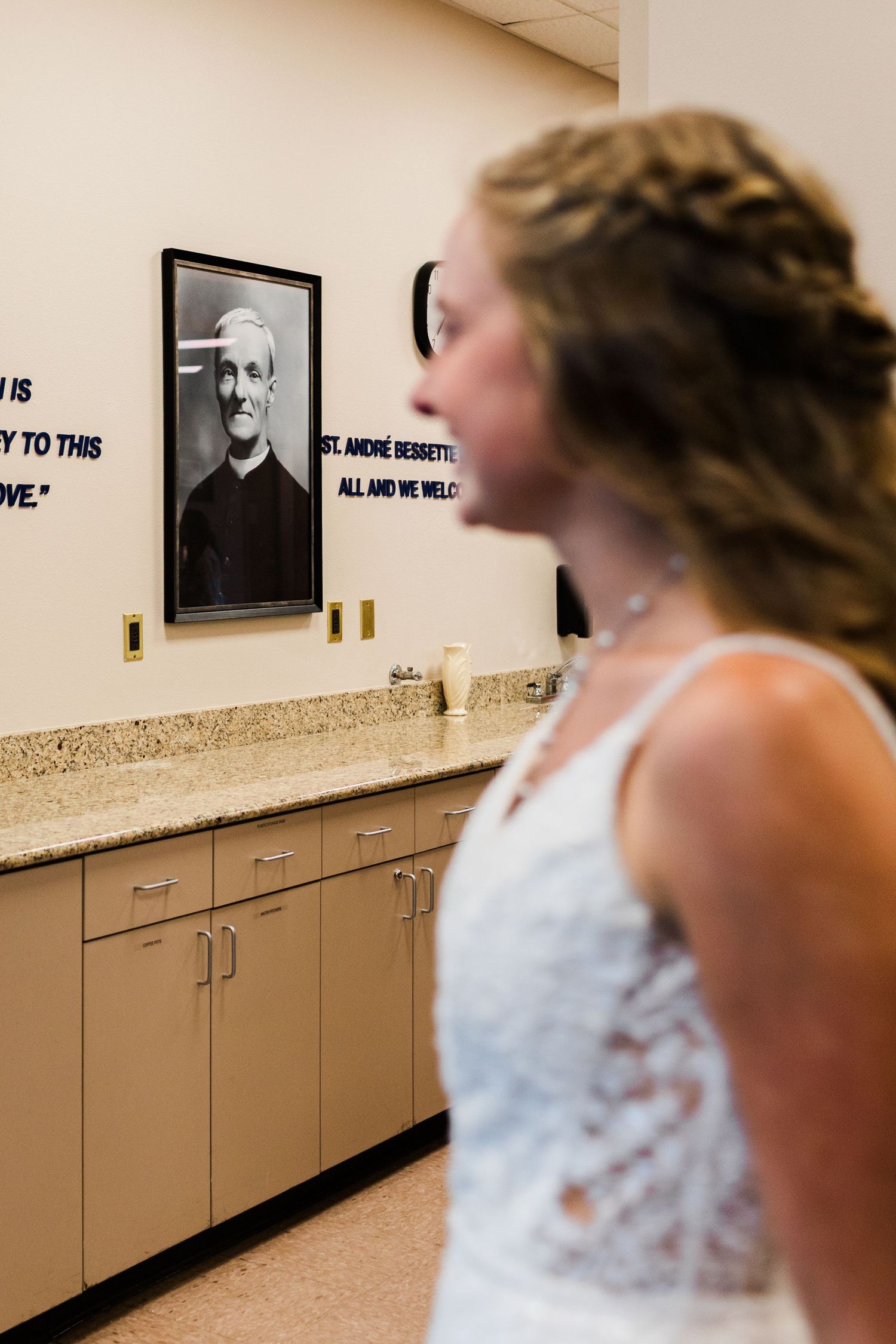 Bride in white lace dress views portrait of St. Andre Bessette on wall above tan cabinets in chapel room.