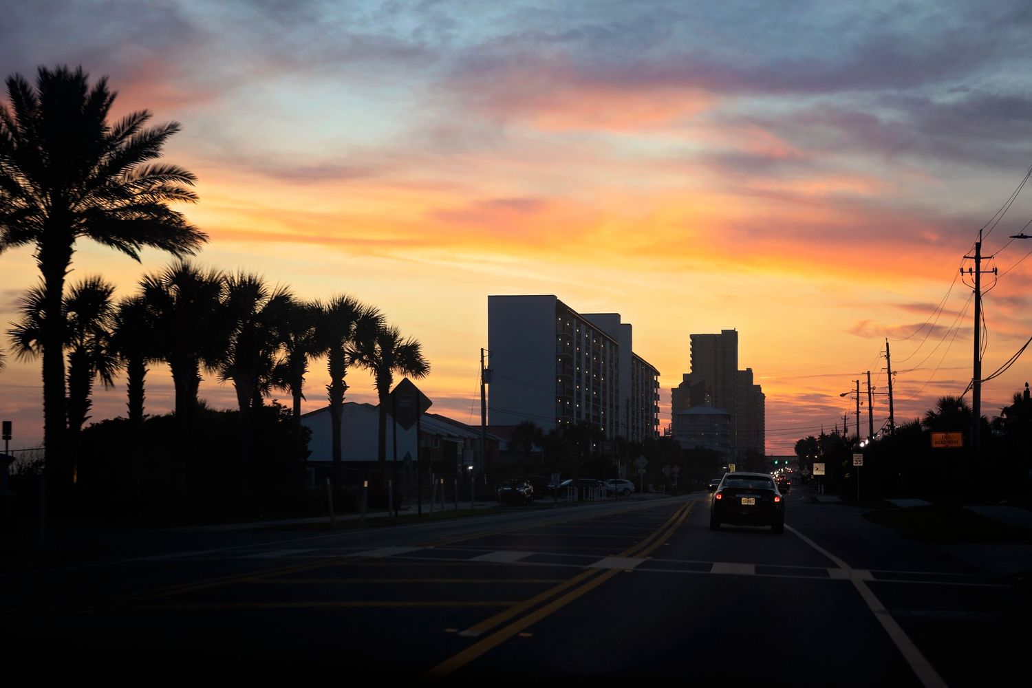 Palm trees and buildings silhouetted against a dramatic orange and purple coastal sunset.