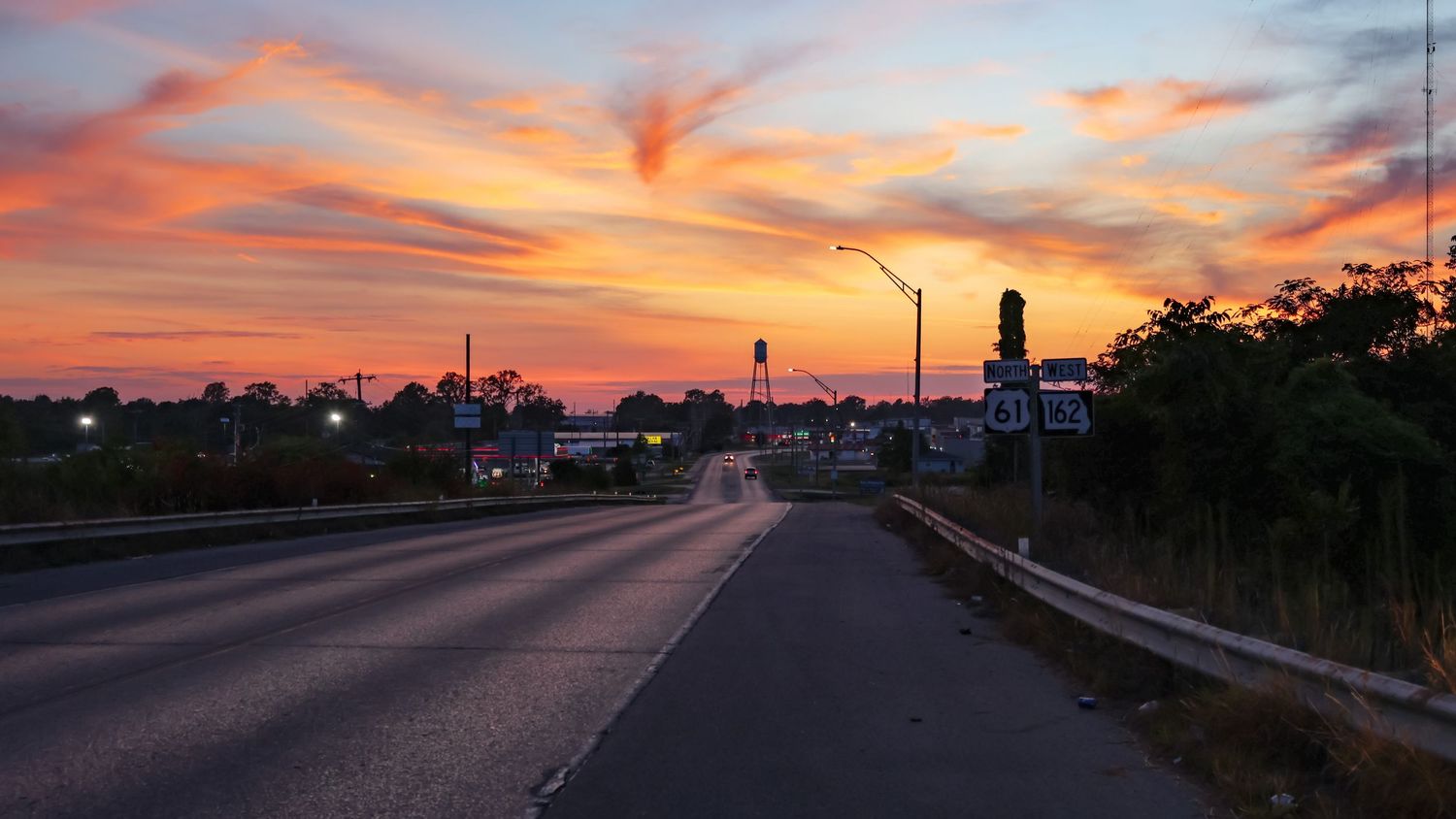 Dramatic orange and pink sunset sky over a highway with street lights and silhouetted buildings.