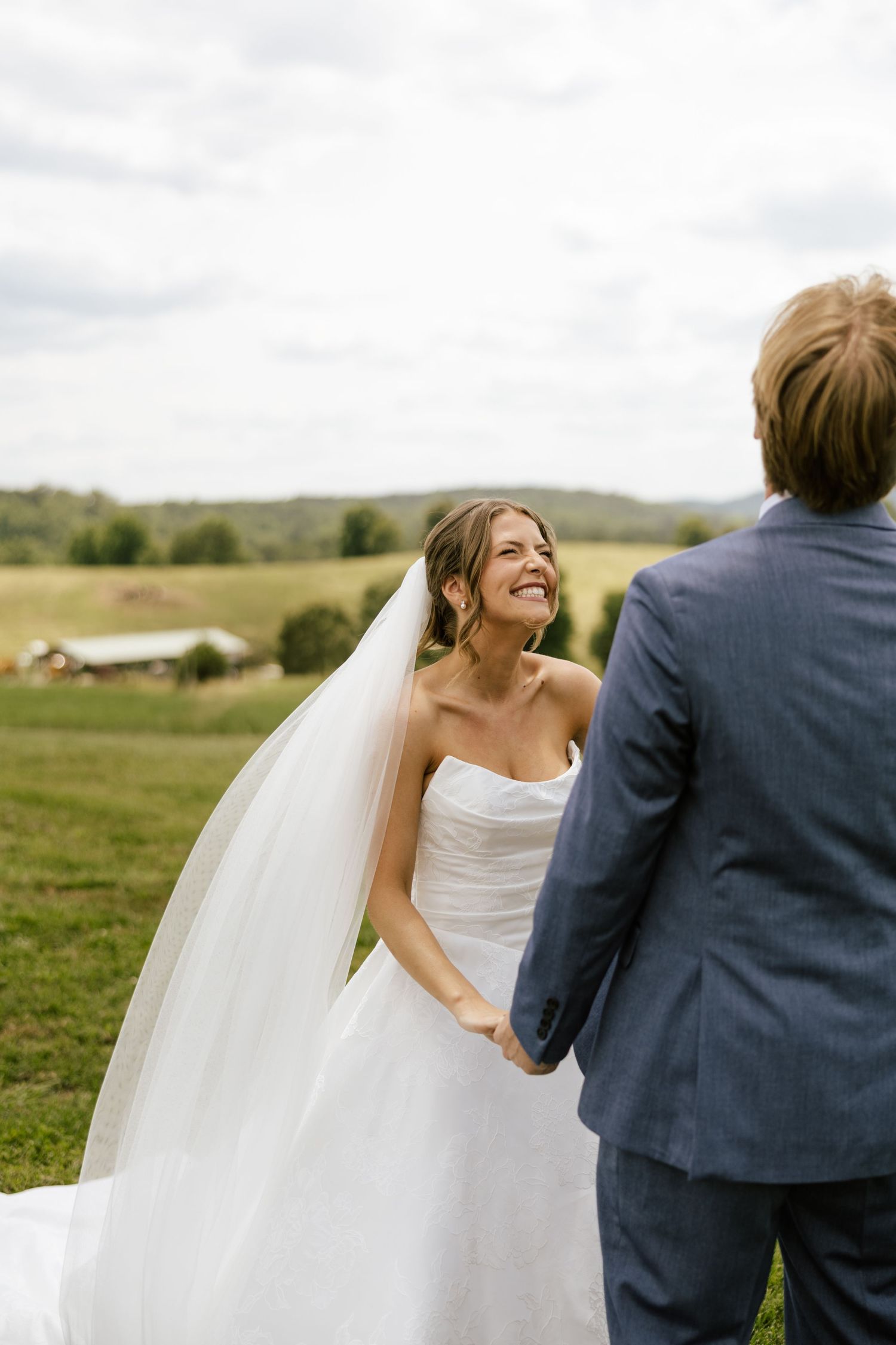 Smiling bride in white strapless gown and veil holds hands with groom in blue suit outdoors on lush green farm.
