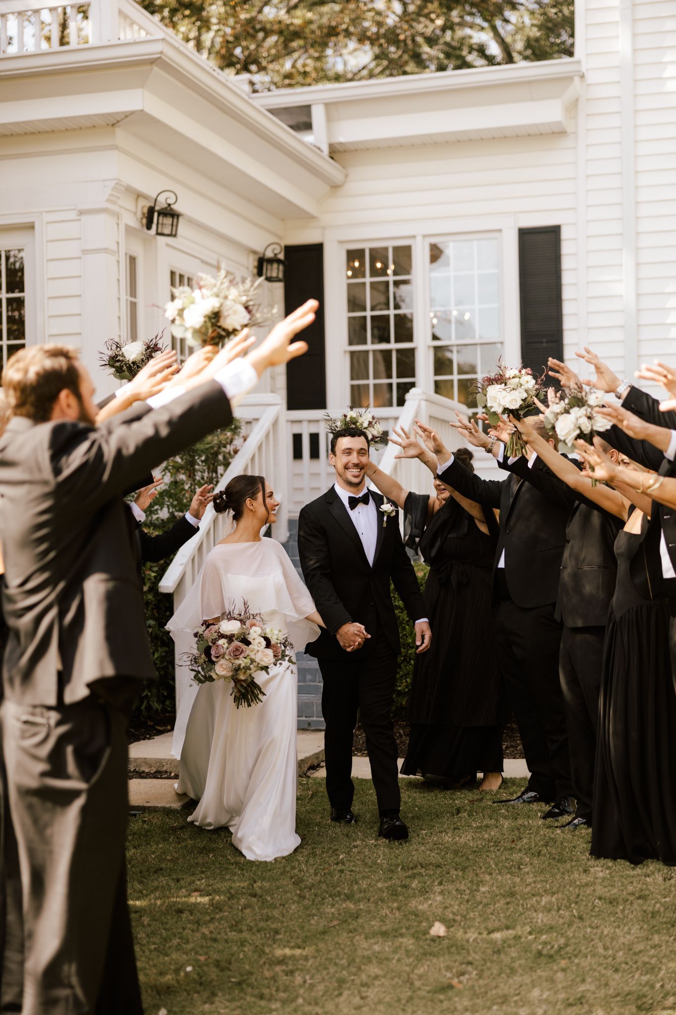 Joyful newlyweds exit ceremony as wedding party raises arms in celebration outside a classic white colonial venue.