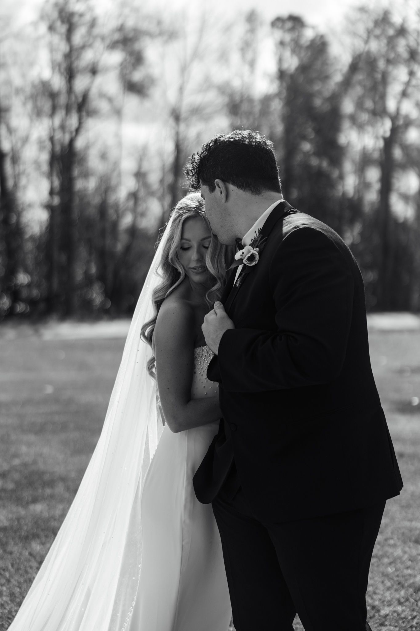 Black and white photo of groom embracing bride in wedding gown and veil outdoors among trees.