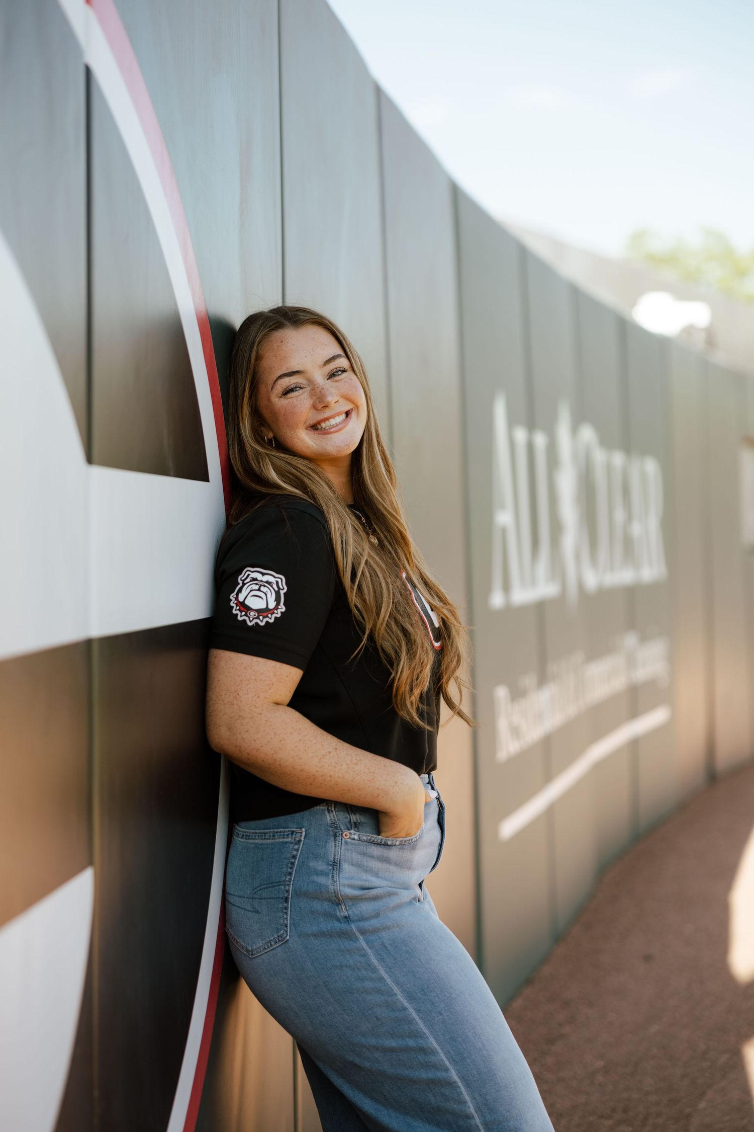 Smiling young woman in black graphic tee and jeans leaning against a wall outdoors in warm sunlight.