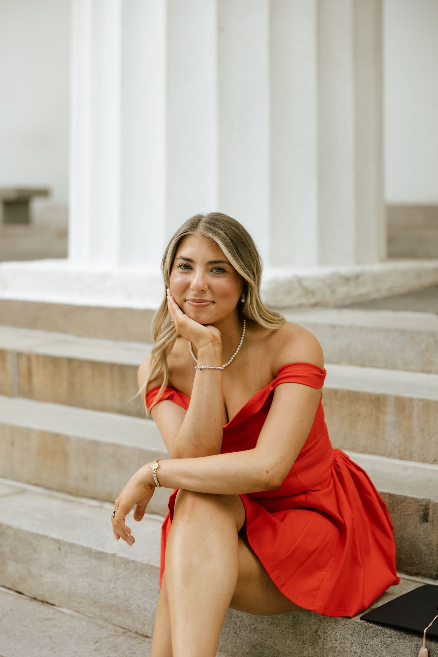Young woman in red off-shoulder dress sitting on marble steps, smiling, wearing pearl necklace and gold bracelet.