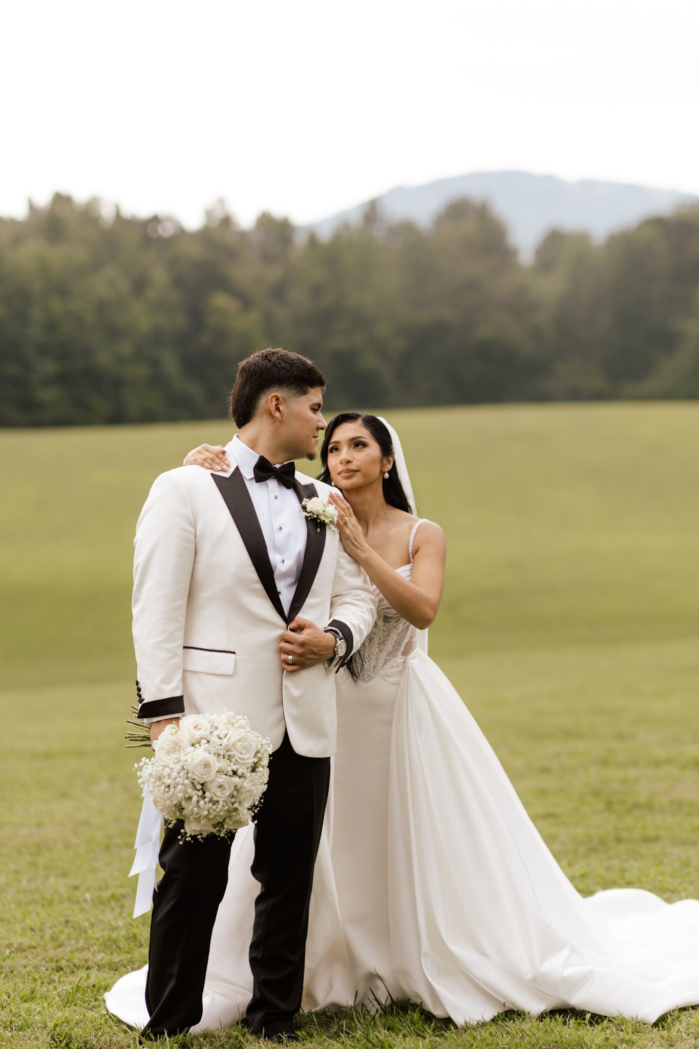 Newlyweds in white attire share tender moments on a grassy field.