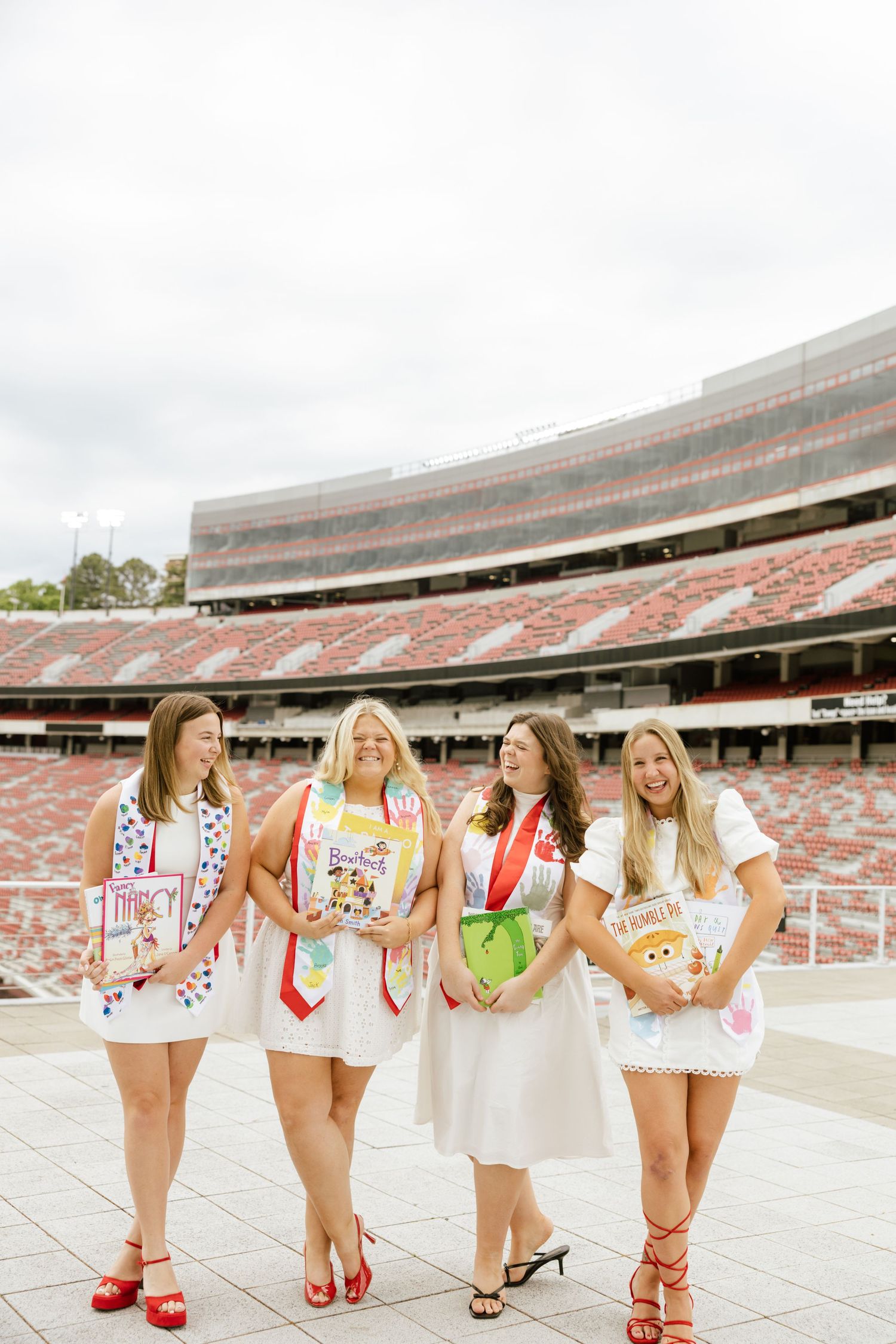 Four female graduates in white dresses with graduation stoles pose together at a large college football stadium.
