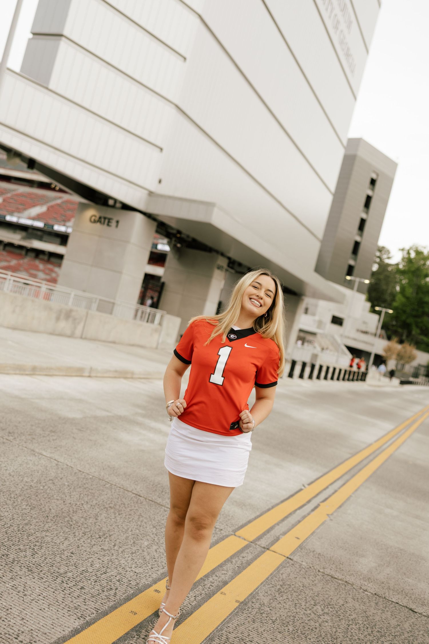 Smiling blonde woman in red Nike football jersey #1 and white skirt posing outside a stadium Gate 1.