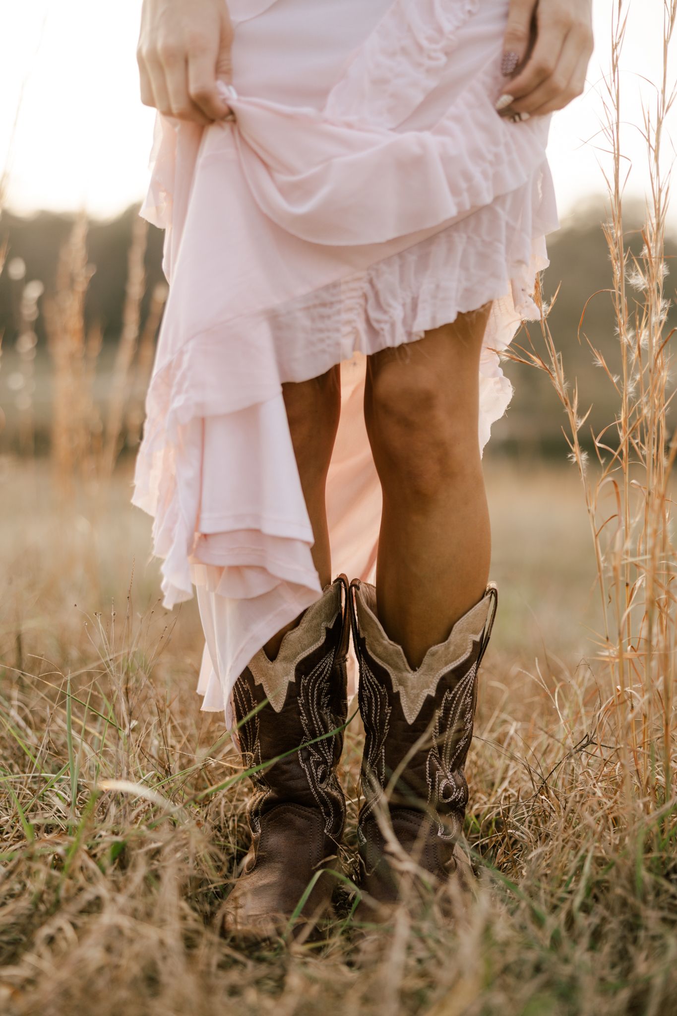 Woman in white ruffled dress and brown cowboy boots standing in golden field at sunset.