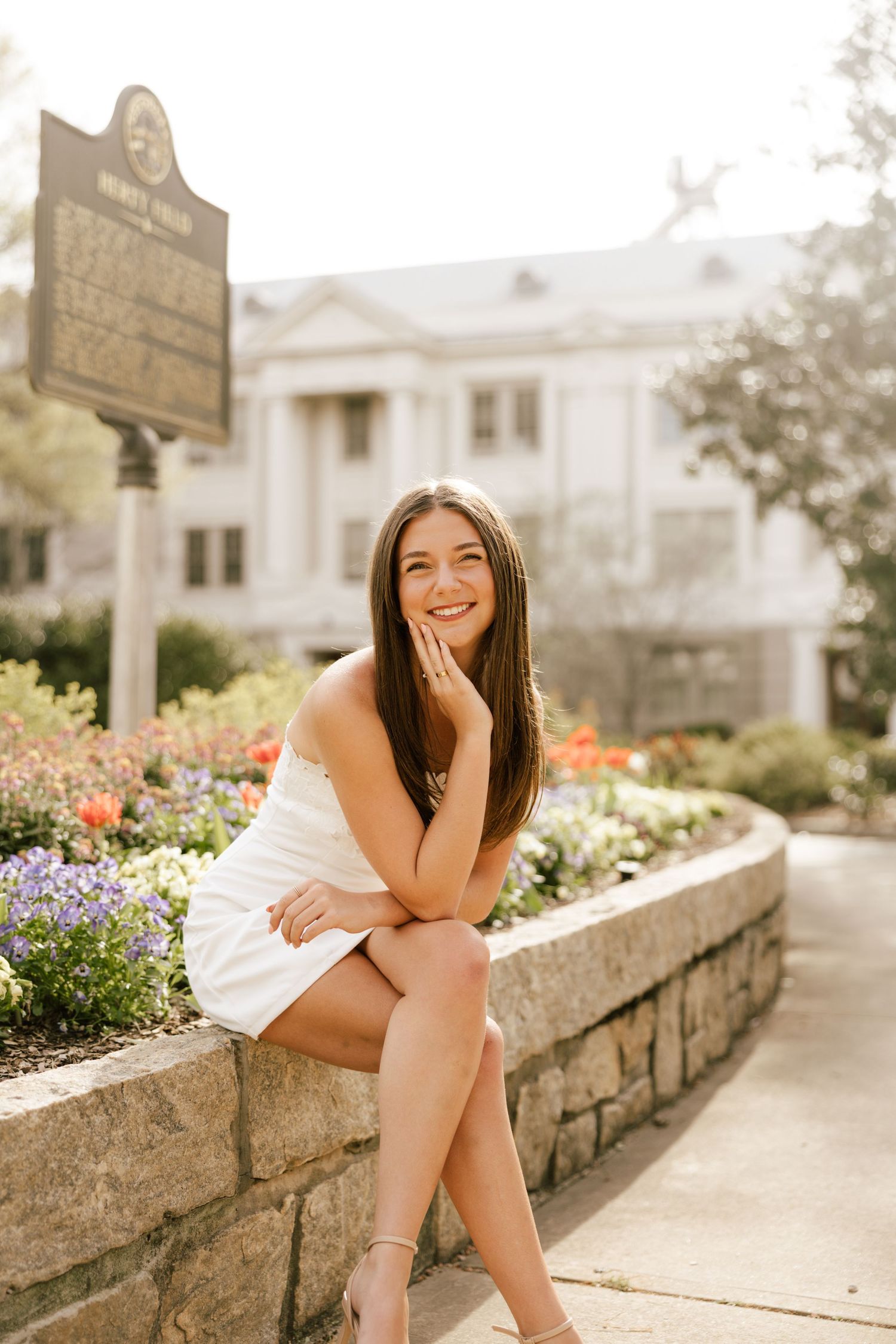 Young woman in white dress sitting on stone wall surrounded by colorful flowers, smiling outdoors near historic building.