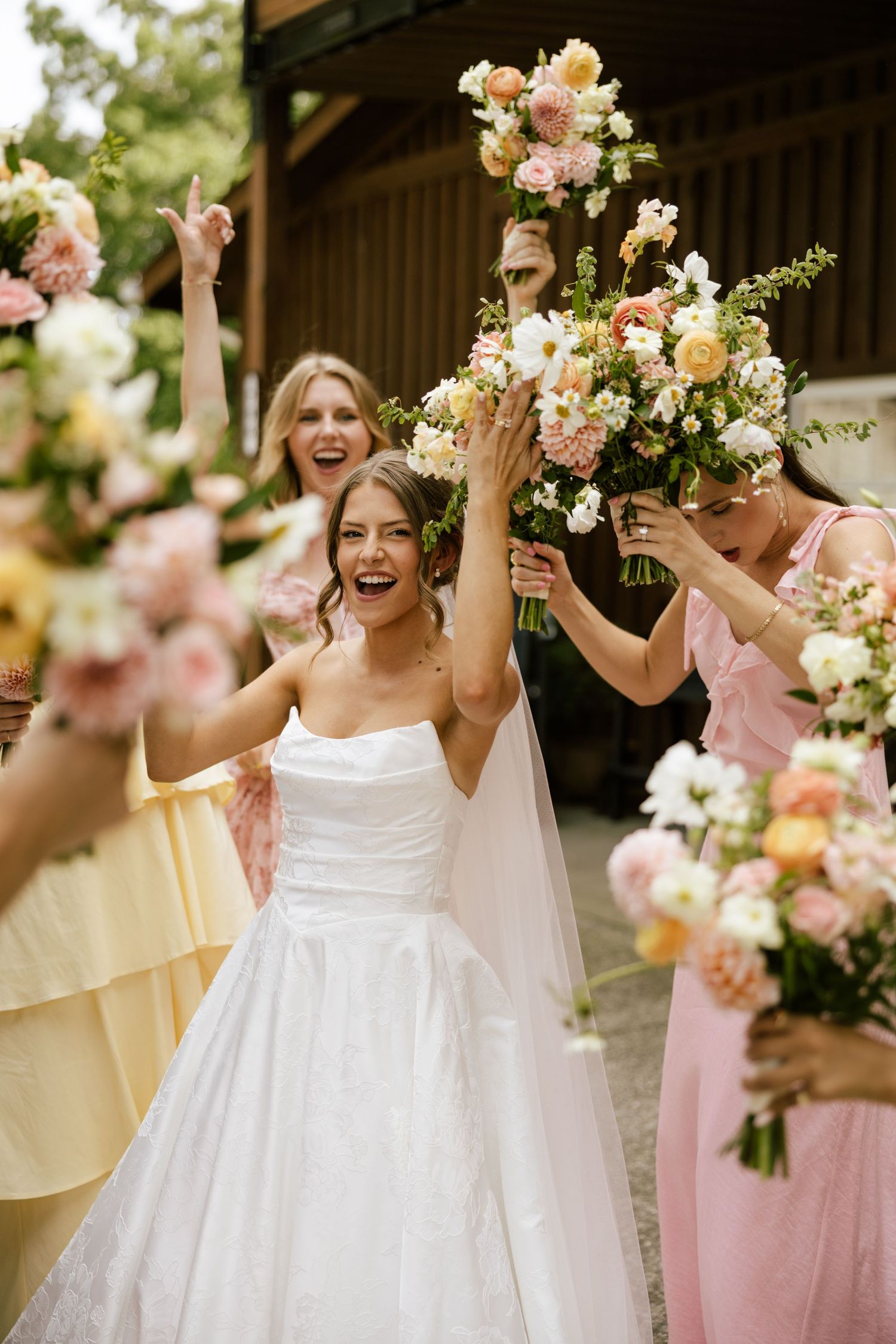 Joyful bride in white gown raises bouquet with bridesmaids in pink and yellow dresses celebrating outdoors.