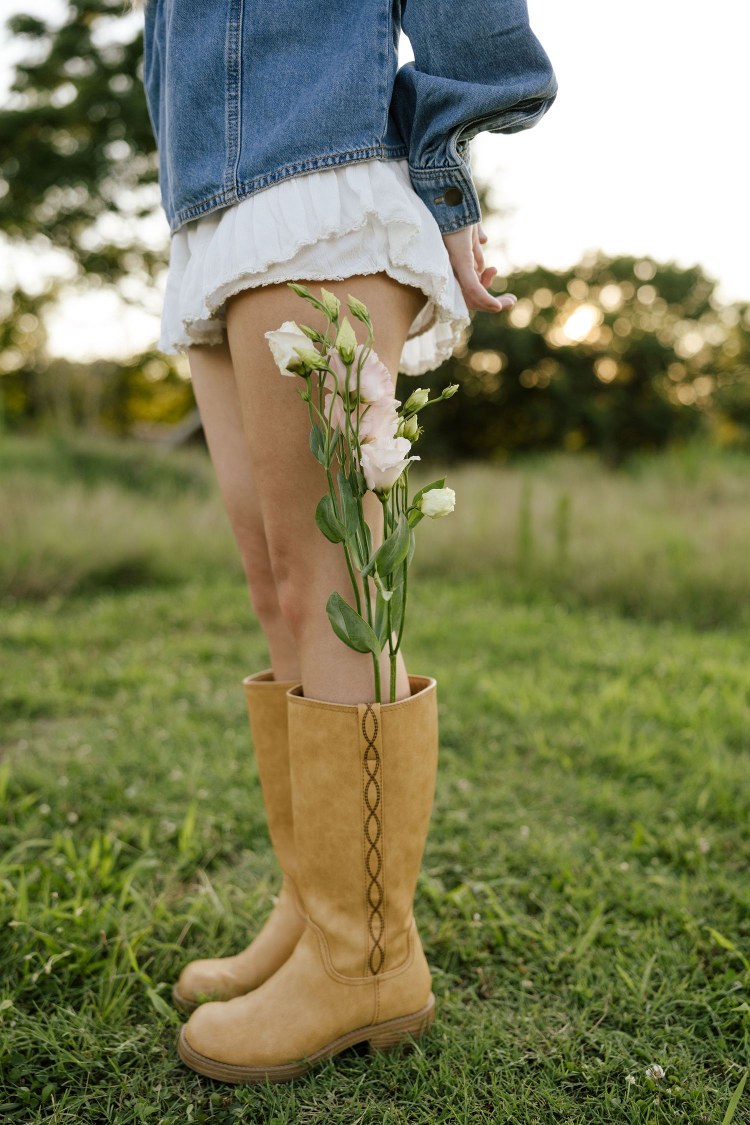Woman in denim jacket and white skirt wearing tan knee-high boots with pink flowers tucked inside, standing on green grass.