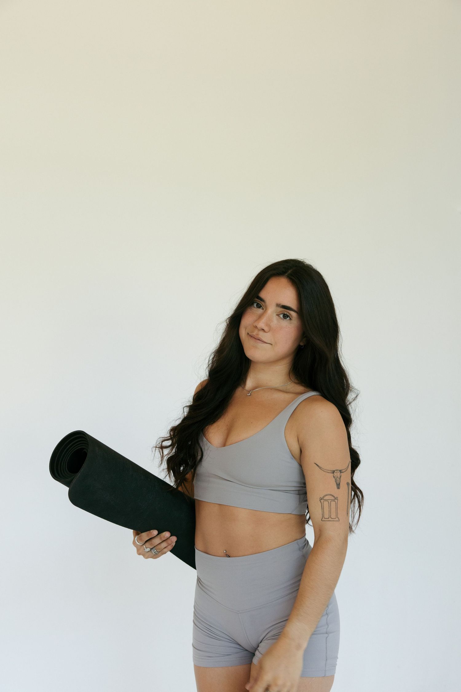 Woman in gray athletic set holding a rolled black yoga mat against white background.