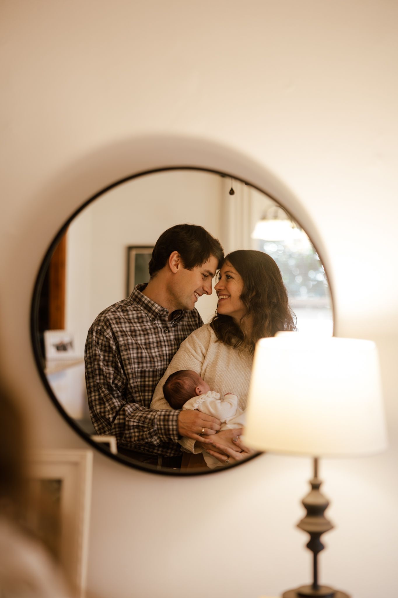 A couple tenderly touches foreheads while holding their newborn baby, reflected in a round mirror with warm lamp light.