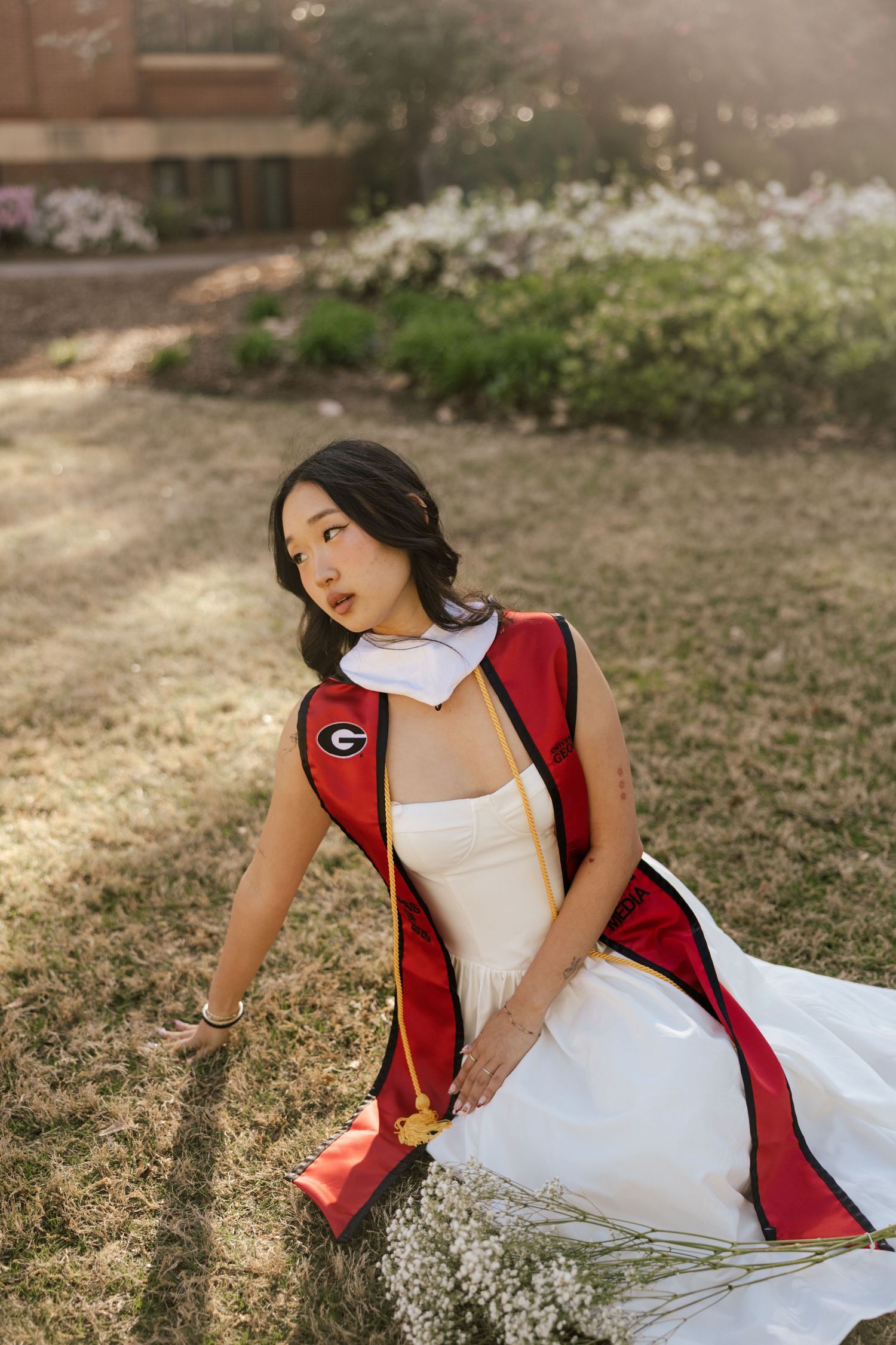 Young woman in white dress and red University of Georgia graduation stole sitting on grass outdoors.