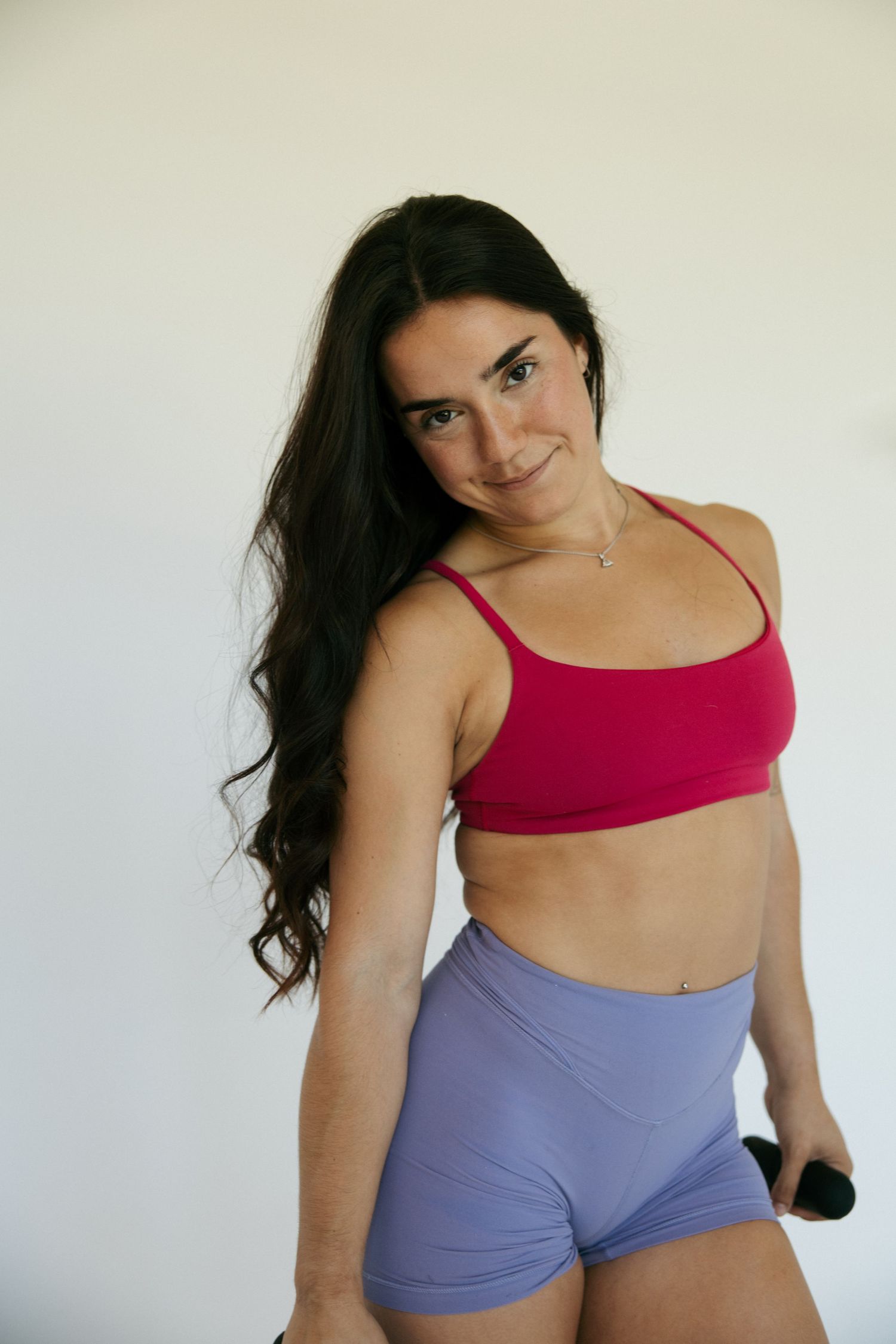 Woman in pink sports bra and lavender shorts holding dumbbell, posing against white background.