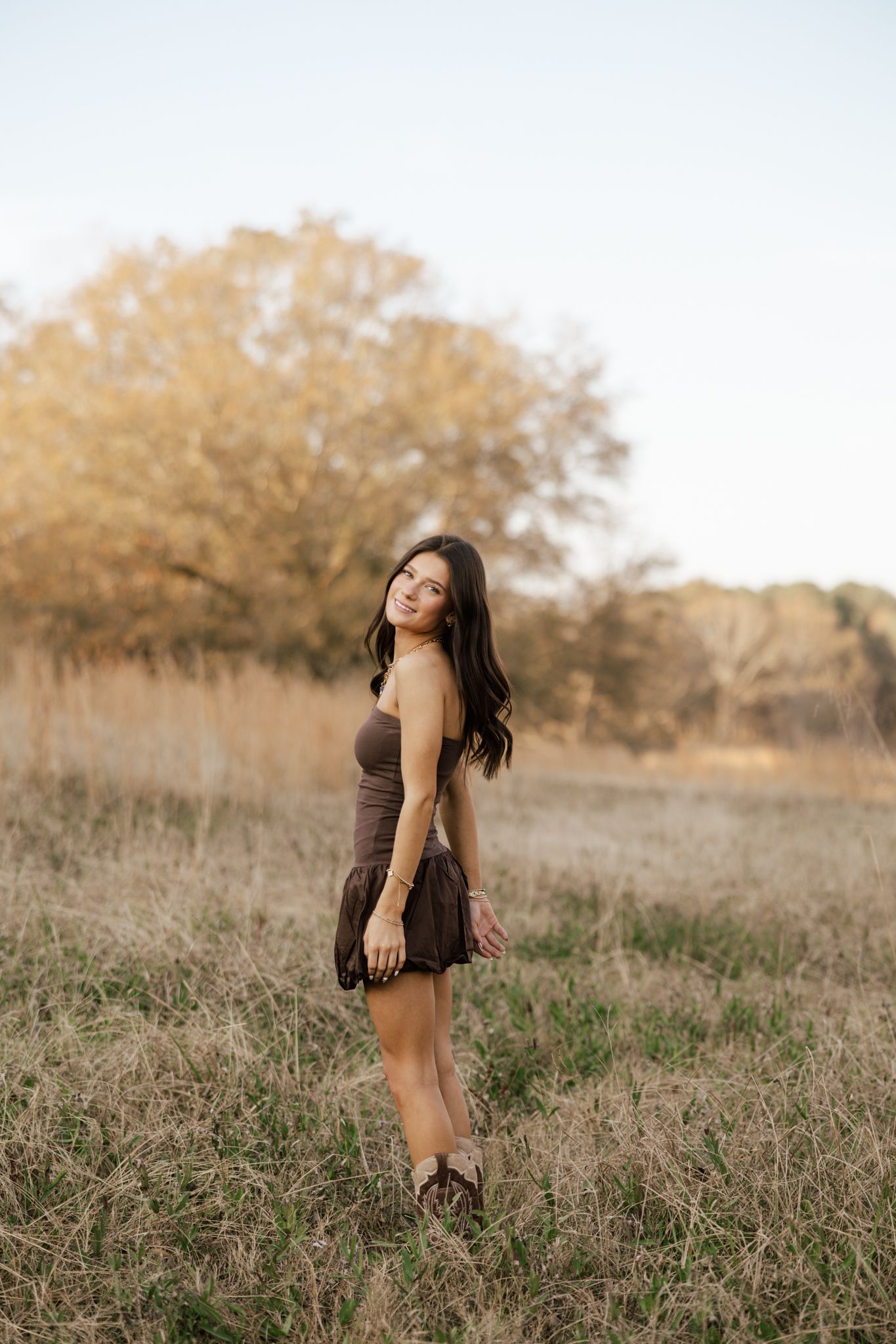 Young woman in brown mini dress standing in golden field at sunset with trees in background.