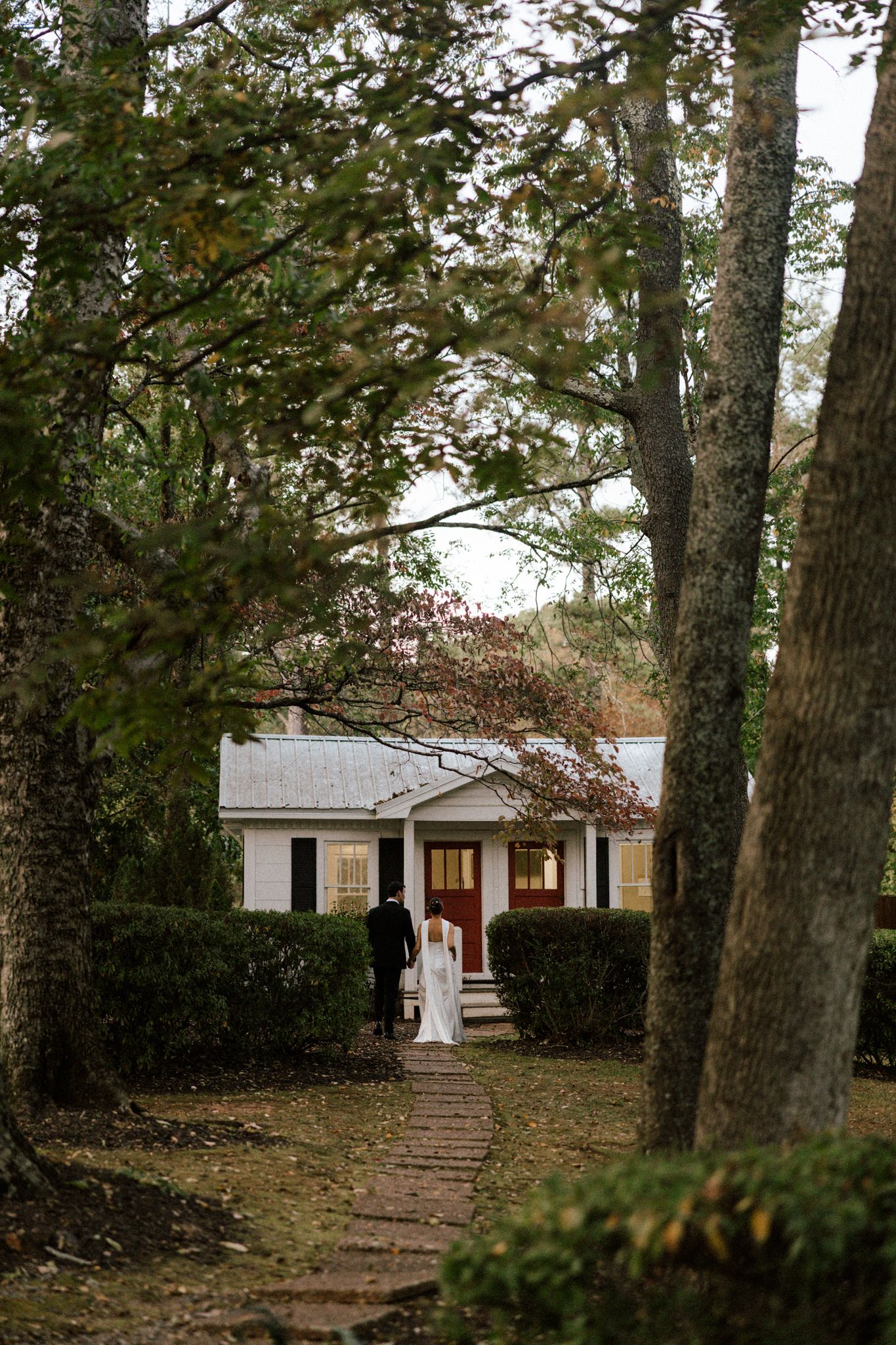 Bride and groom walk hand-in-hand along a brick path toward a charming white cottage surrounded by lush trees.