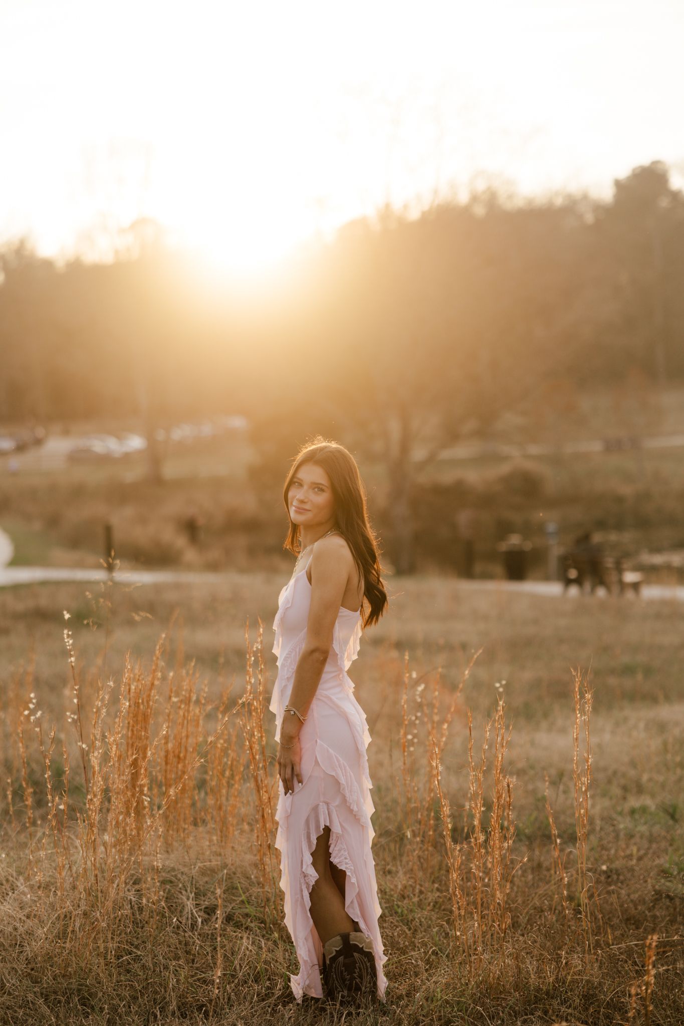 A young woman in a flowing pink ruffled dress stands in a golden field during a stunning sunset.