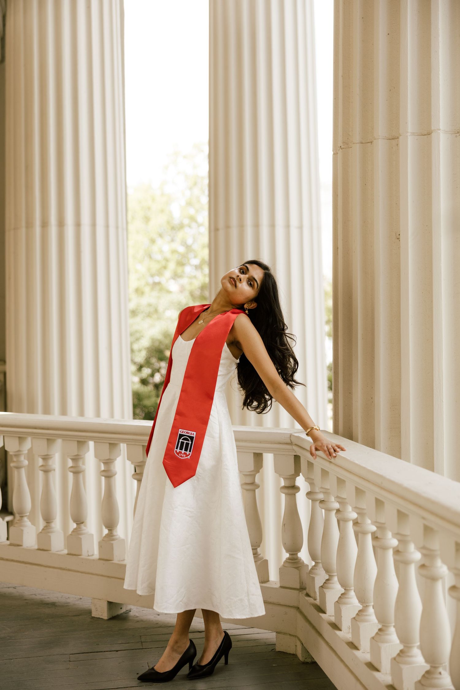 Graduate in white dress and red academic stole poses gracefully by white columns and balustrade.