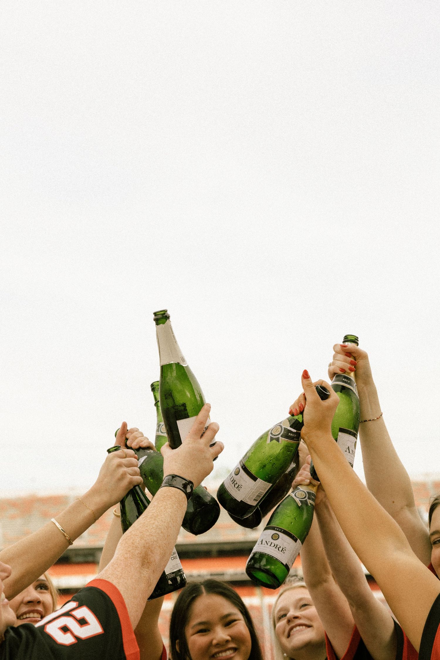 Group of young people celebrating outdoors, raising green champagne bottles in a toast at a sports stadium.
