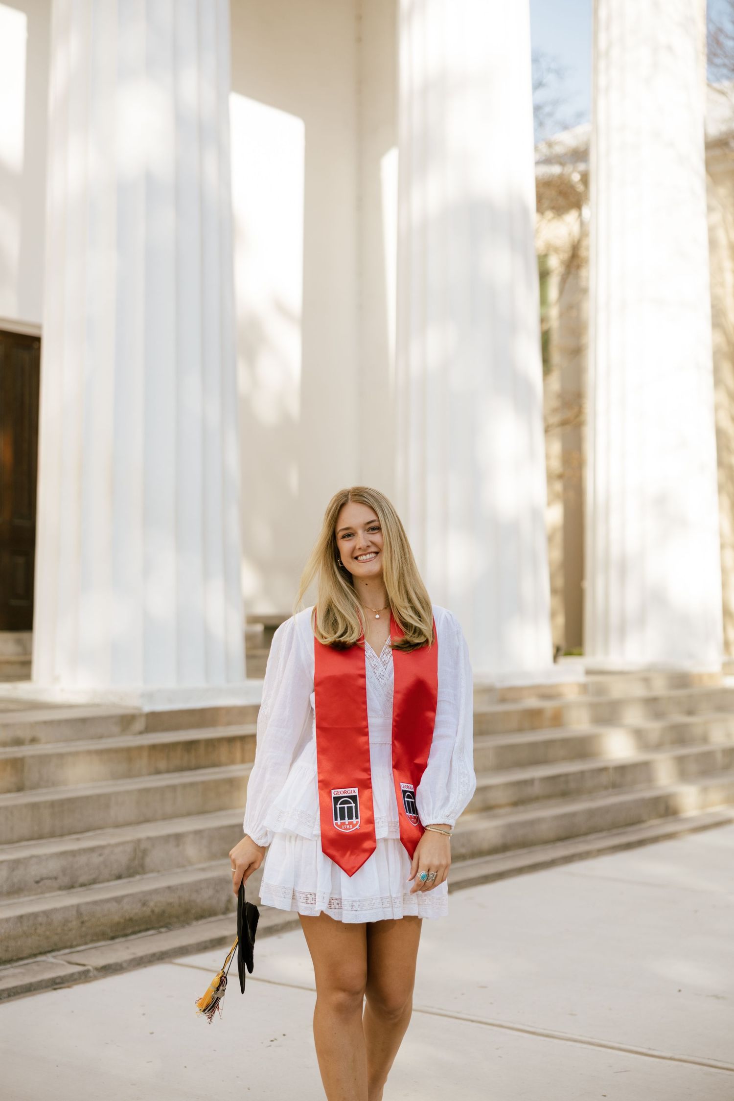 Smiling blonde graduate in white dress with red graduation stole holds cap outside columned building.