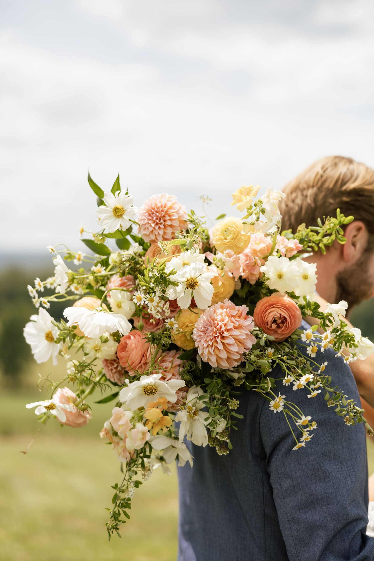 Person holding a large bridal bouquet of peach dahlias, white cosmos, yellow ranunculus, and wildflowers outdoors.