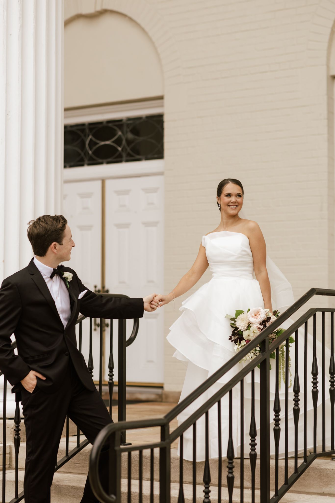 Wedding couple playfully interacting on white columned porch with black railings.