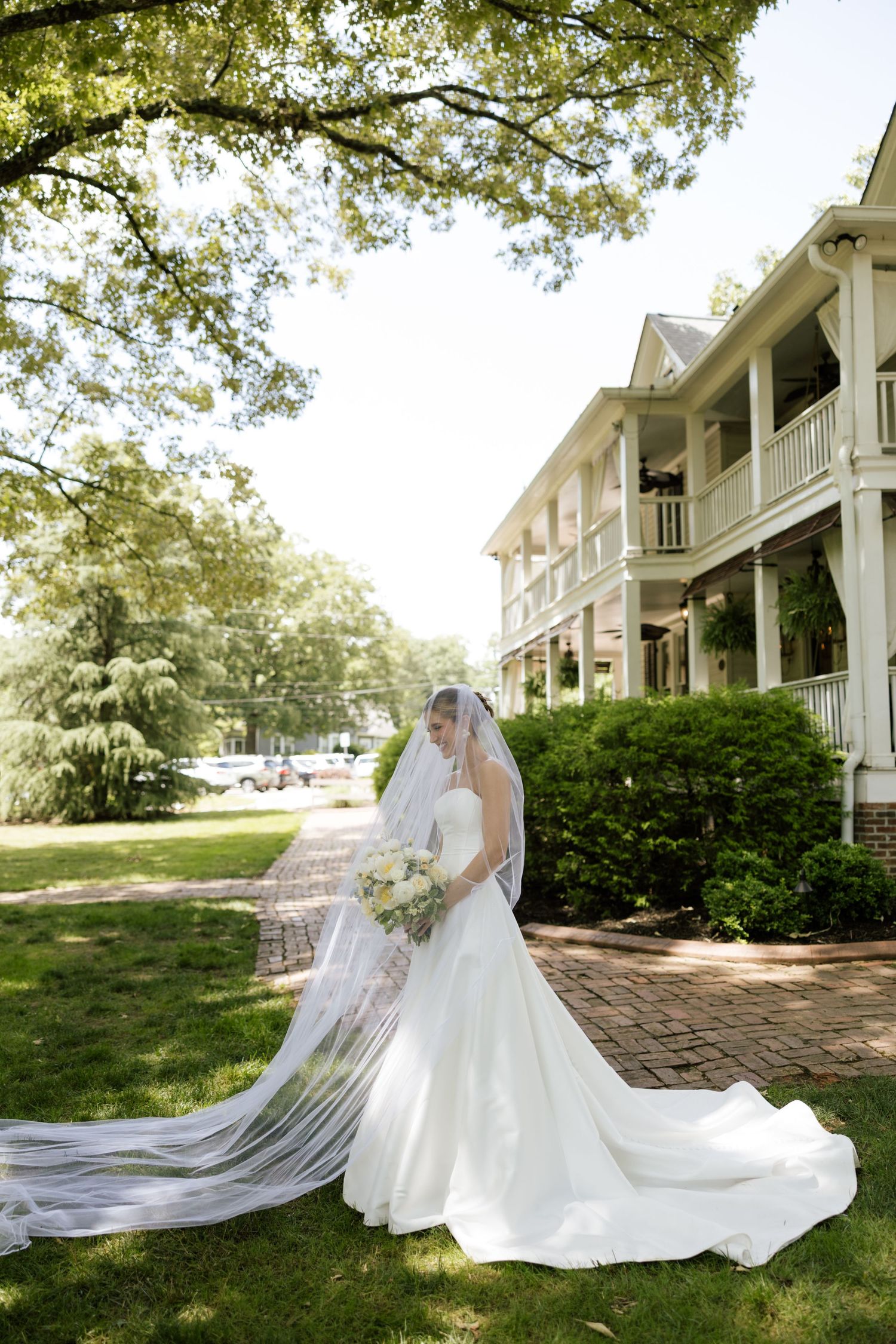 Bride in elegant white gown with long veil holds bouquet outside historic Southern venue.