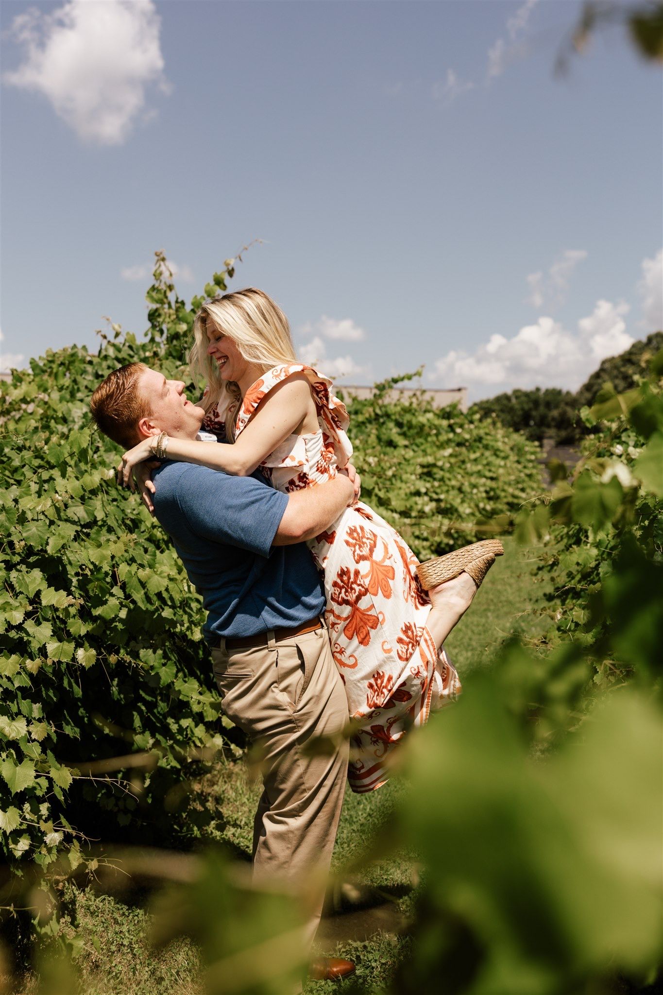Playful couple embraces in a sunny vineyard surrounded by green grapevines.