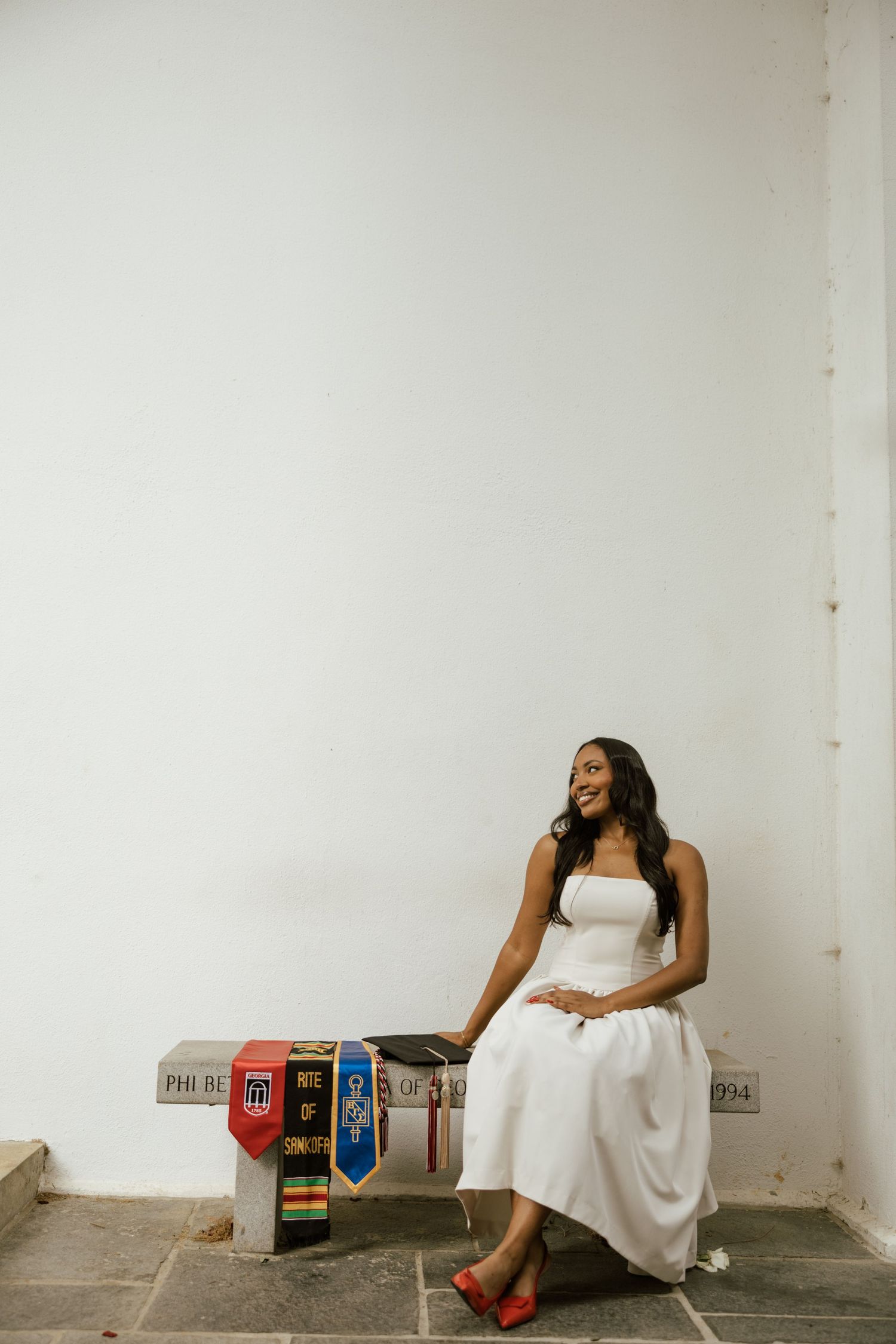 A smiling graduate in a white dress and red heels sits on a stone bench with colorful academic stoles beside her.