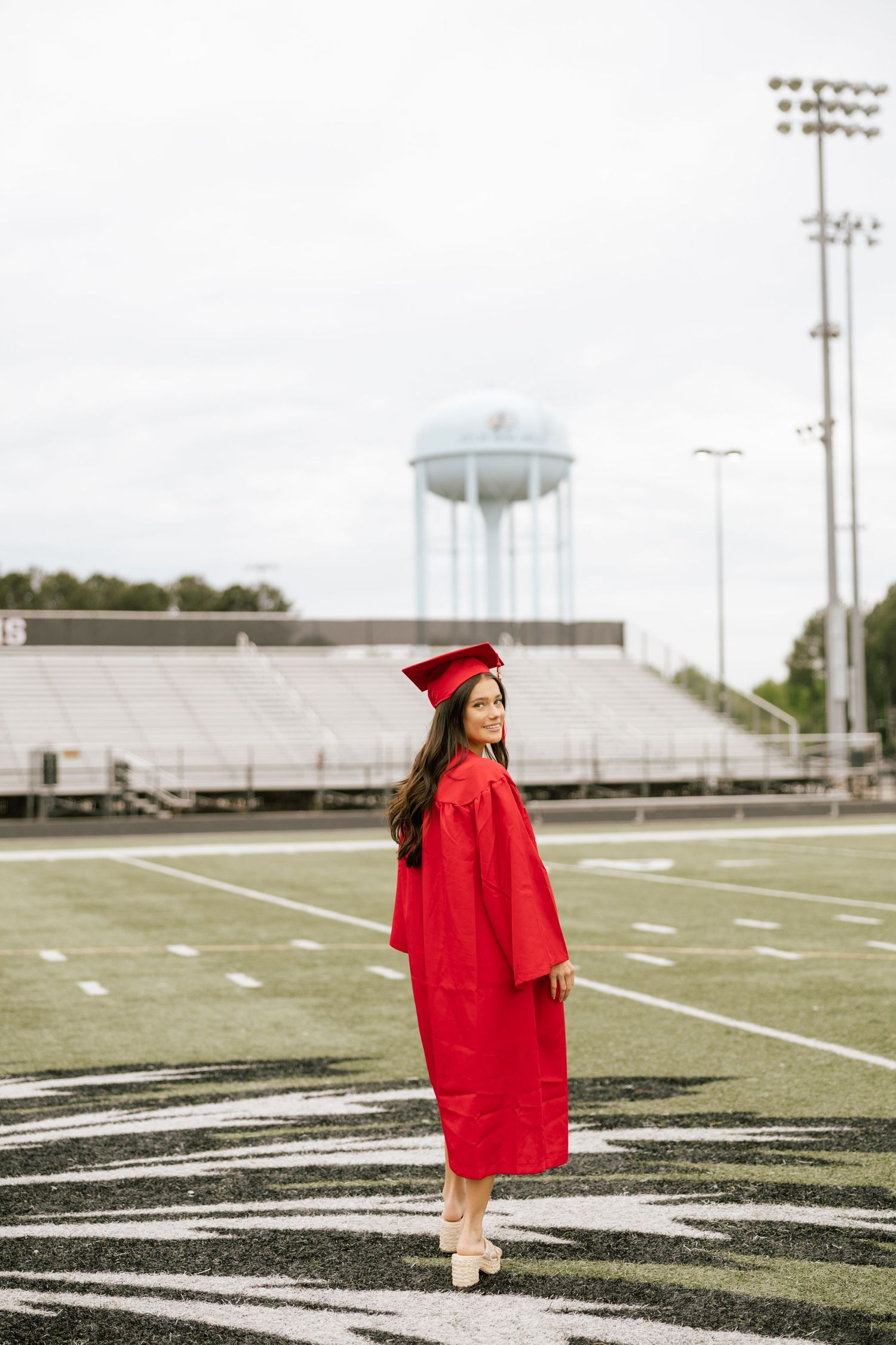 Young woman in red graduation cap and gown standing on football field with stadium bleachers and water tower behind her.