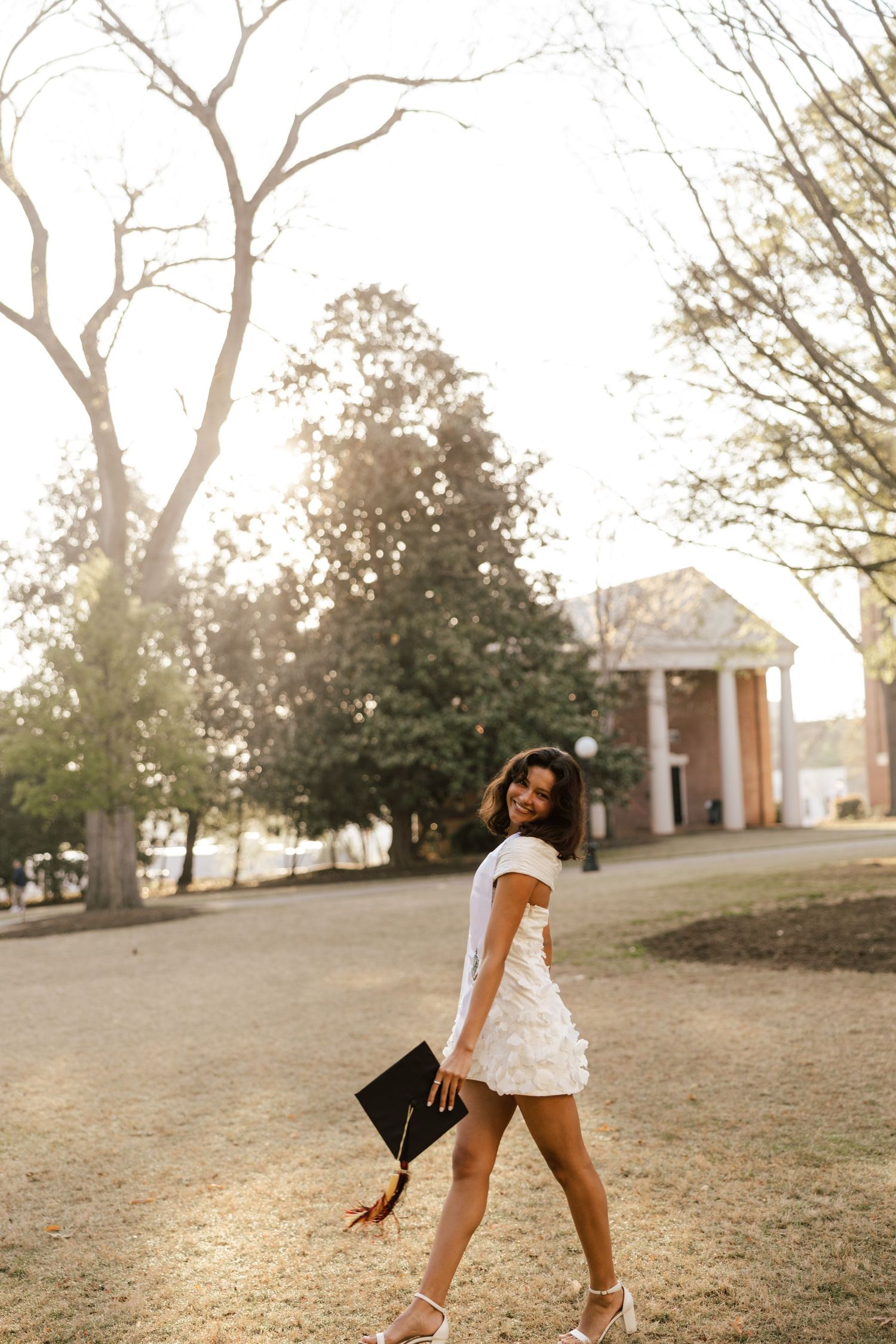 Young woman in white dress holding graduation cap, walking outdoors on campus grounds during golden hour.