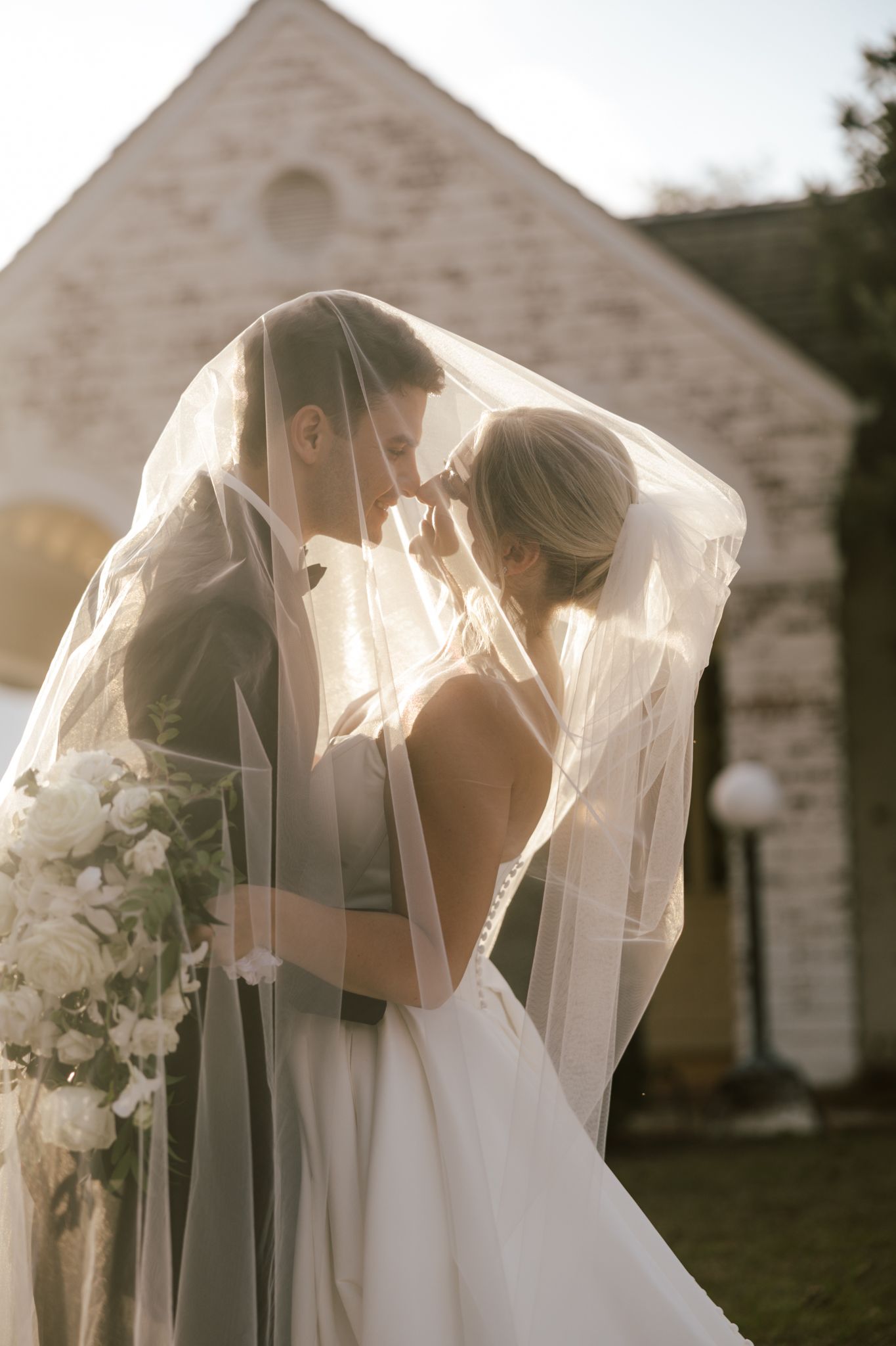 Bride and groom sharing an intimate moment under a flowing veil outside a stone church on their wedding day.