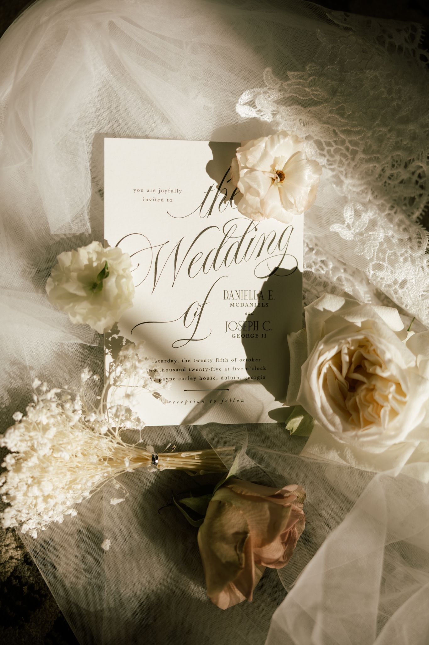Elegant wedding invitation with gold calligraphy surrounded by white roses and tulle fabric in soft natural light.
