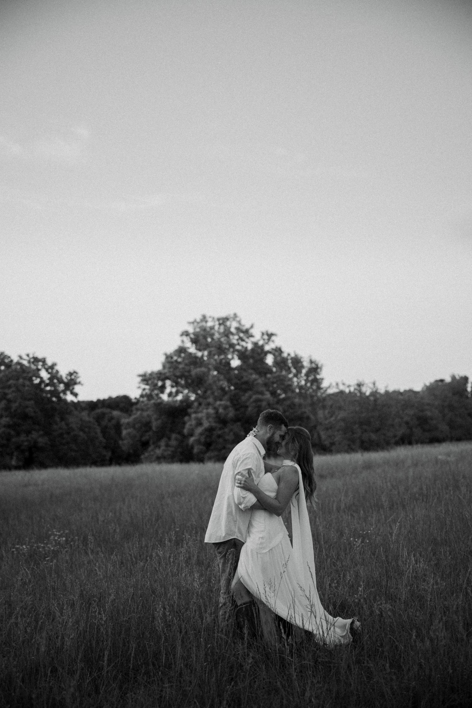 Black and white photo of a couple embracing and kissing in a lush green field with trees in the background.