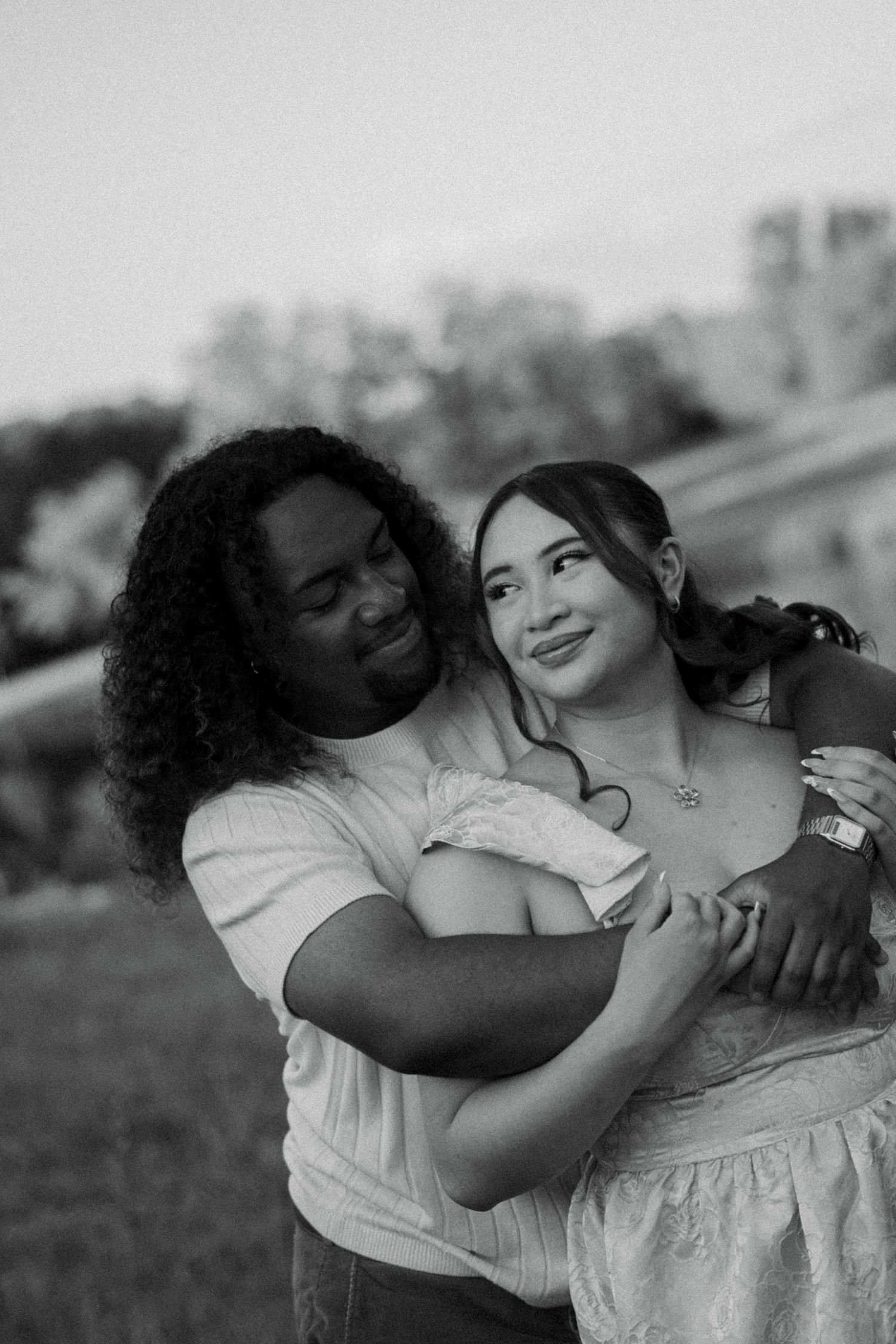 Black and white photo of a couple embracing outdoors, woman smiling as man hugs her from behind.