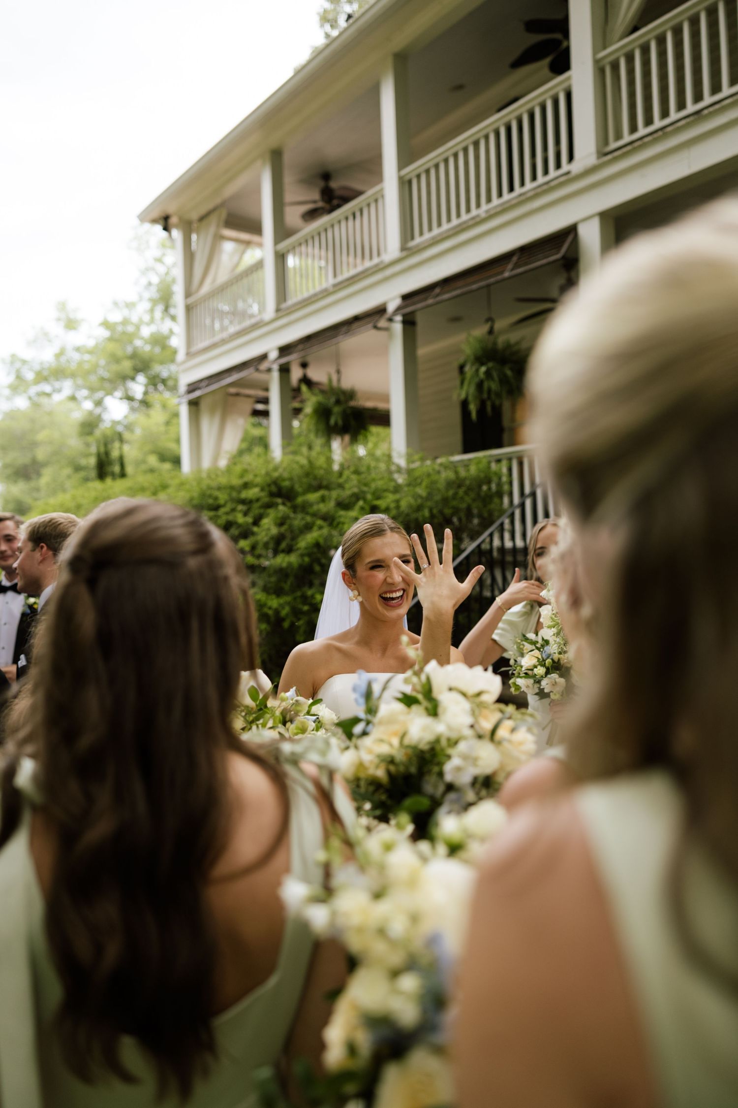 Joyful bride showing her ring to bridesmaids holding white bouquets outside a historic Southern home.