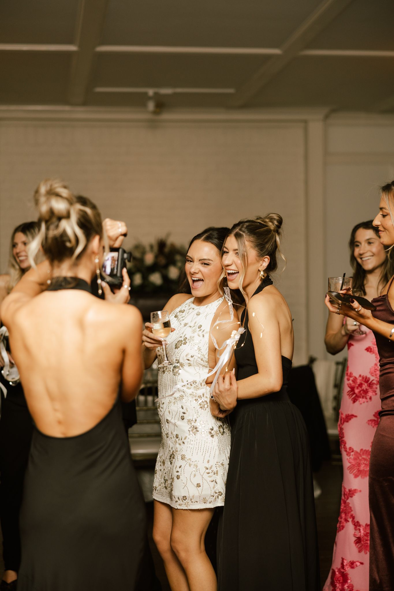 Women in elegant dresses celebrate at a wedding reception, dancing and laughing while holding champagne glasses.