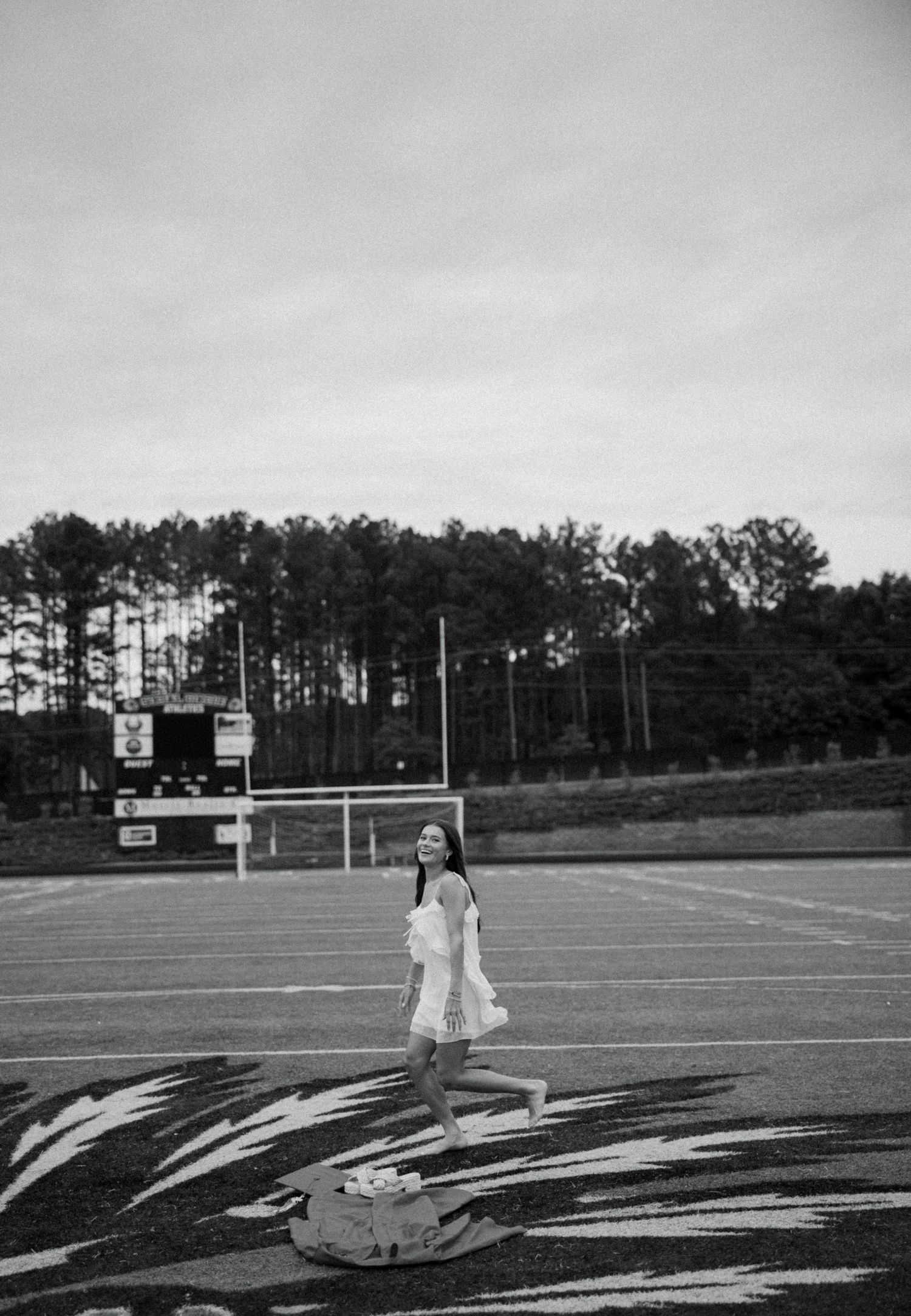 Black and white photo of a young woman in a white dress posing on a football field with trees in the background.