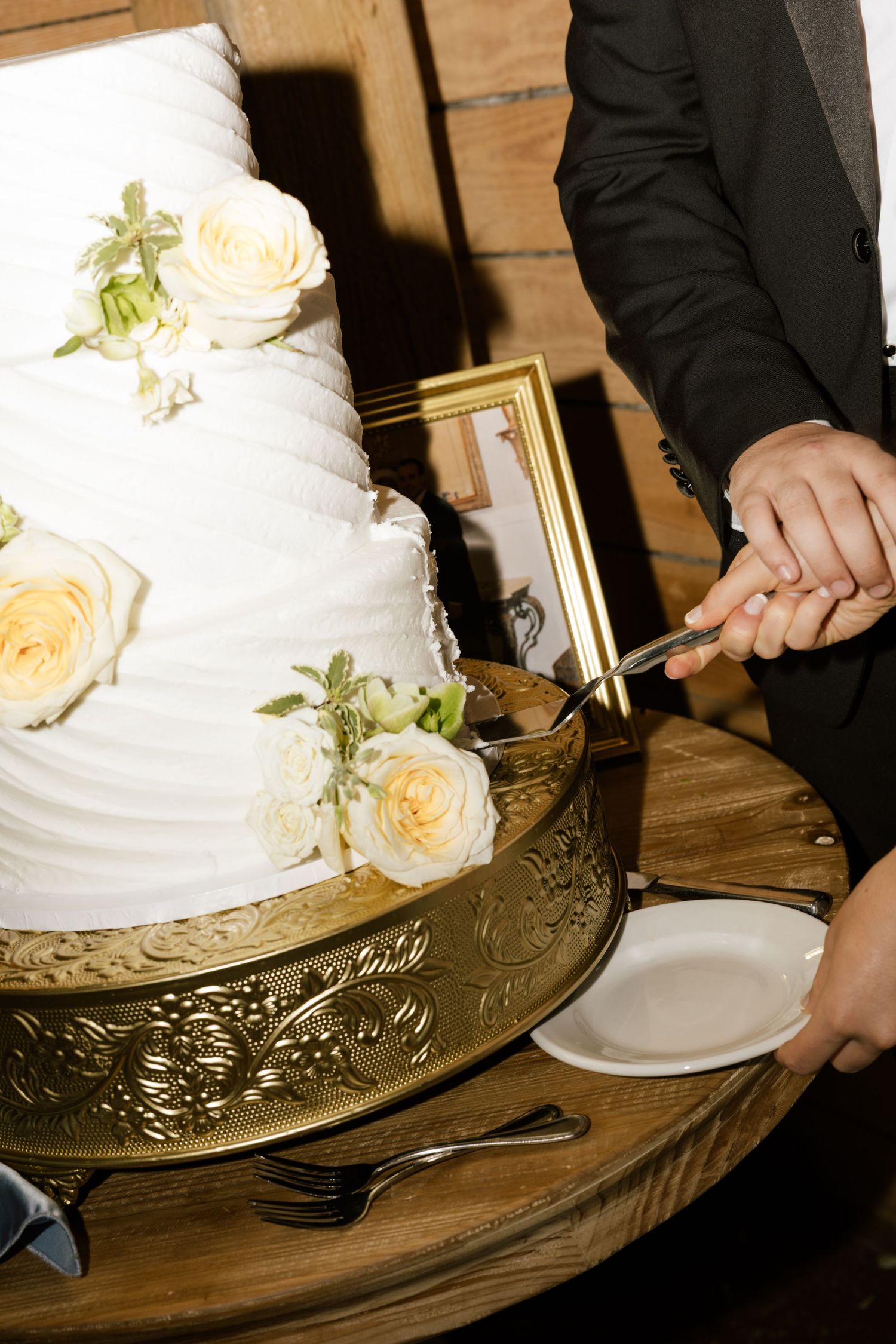 Bride and groom cutting a white three-tier wedding cake with cream roses on a gold ornate cake stand.