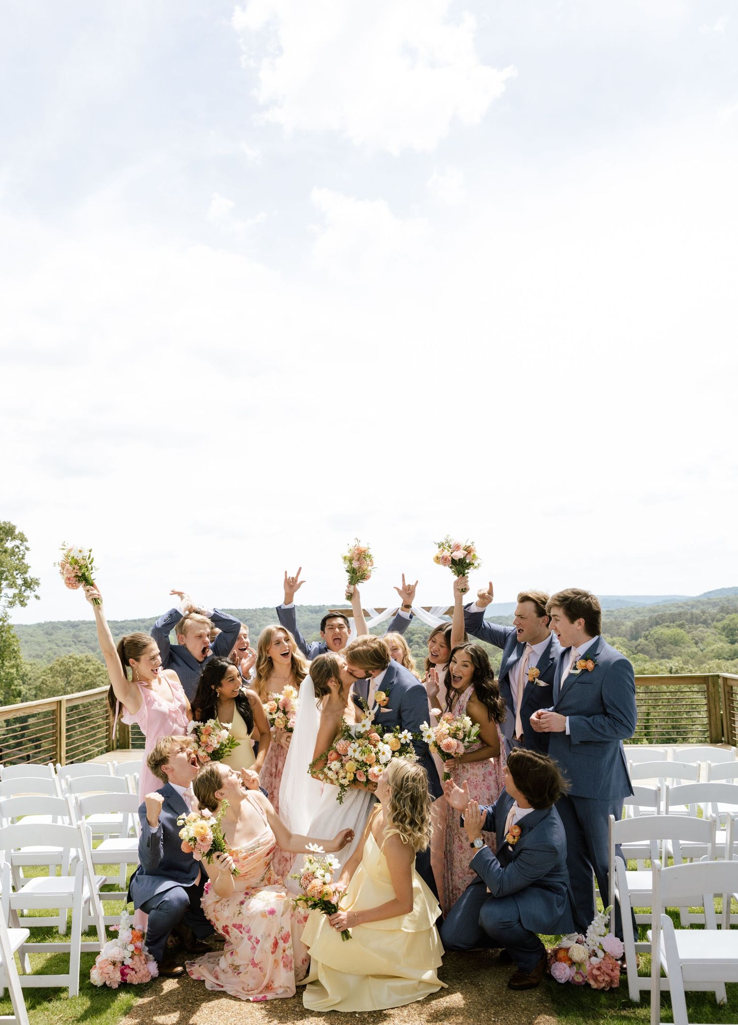 Wedding party celebrating outdoors with bride and groom kissing, bridesmaids in floral dresses, groomsmen in navy suits, bouquets raised.