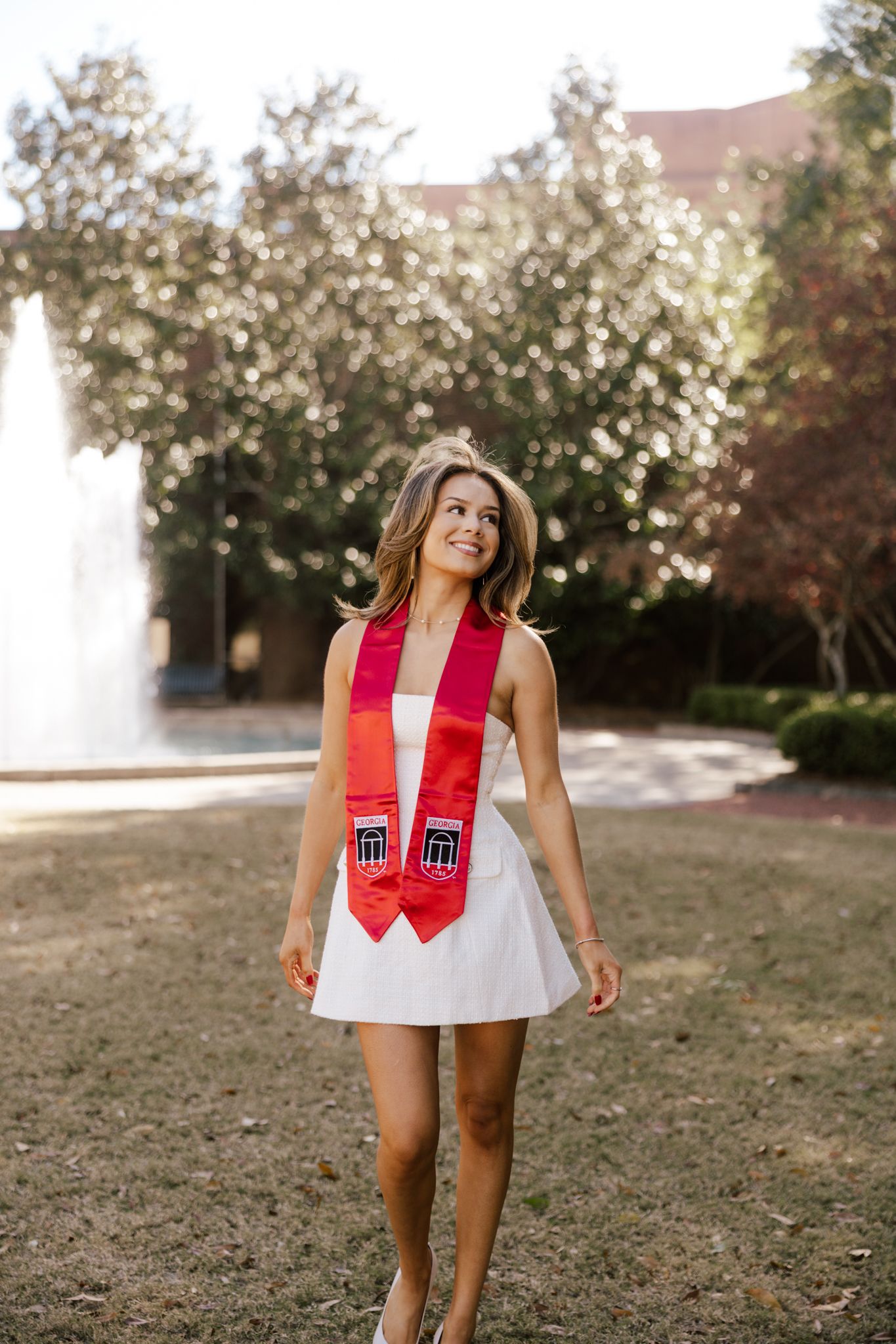 Graduate in white dress and red stole poses near fountain on sunny campus grounds.